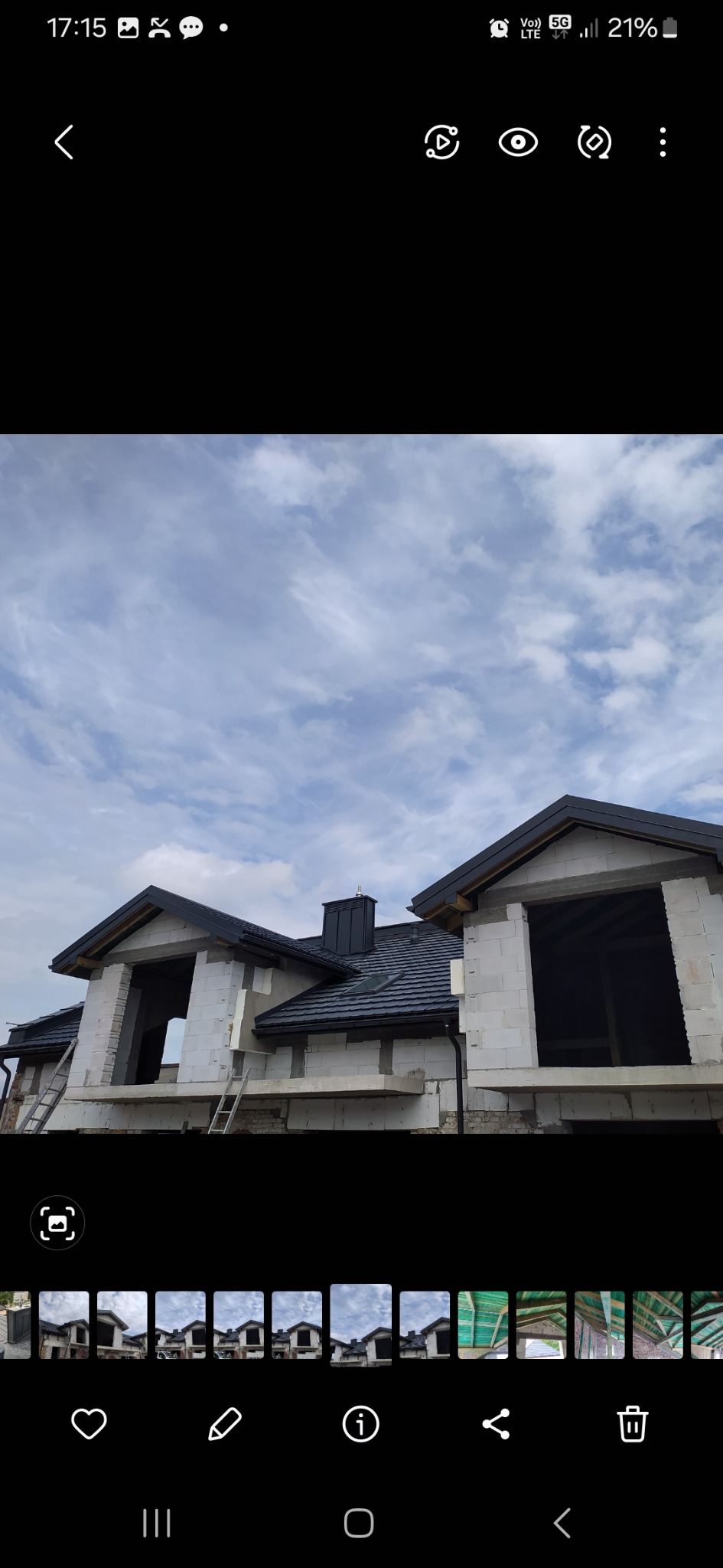 Budowa domu z białych bloczków, widoczny dach z czarną dachówką i komin. Na ścianie drabina. Niebo z chmurami w tle.