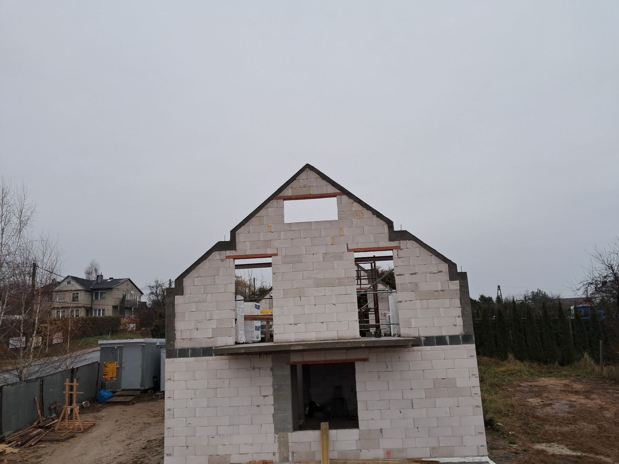 Surowy stan domu z białych bloczków, widoczne otwory okienne i drzwiowe, szare niebo w tle, budowa w toku, widok z dołu, dom w trakcie budowy na działce.