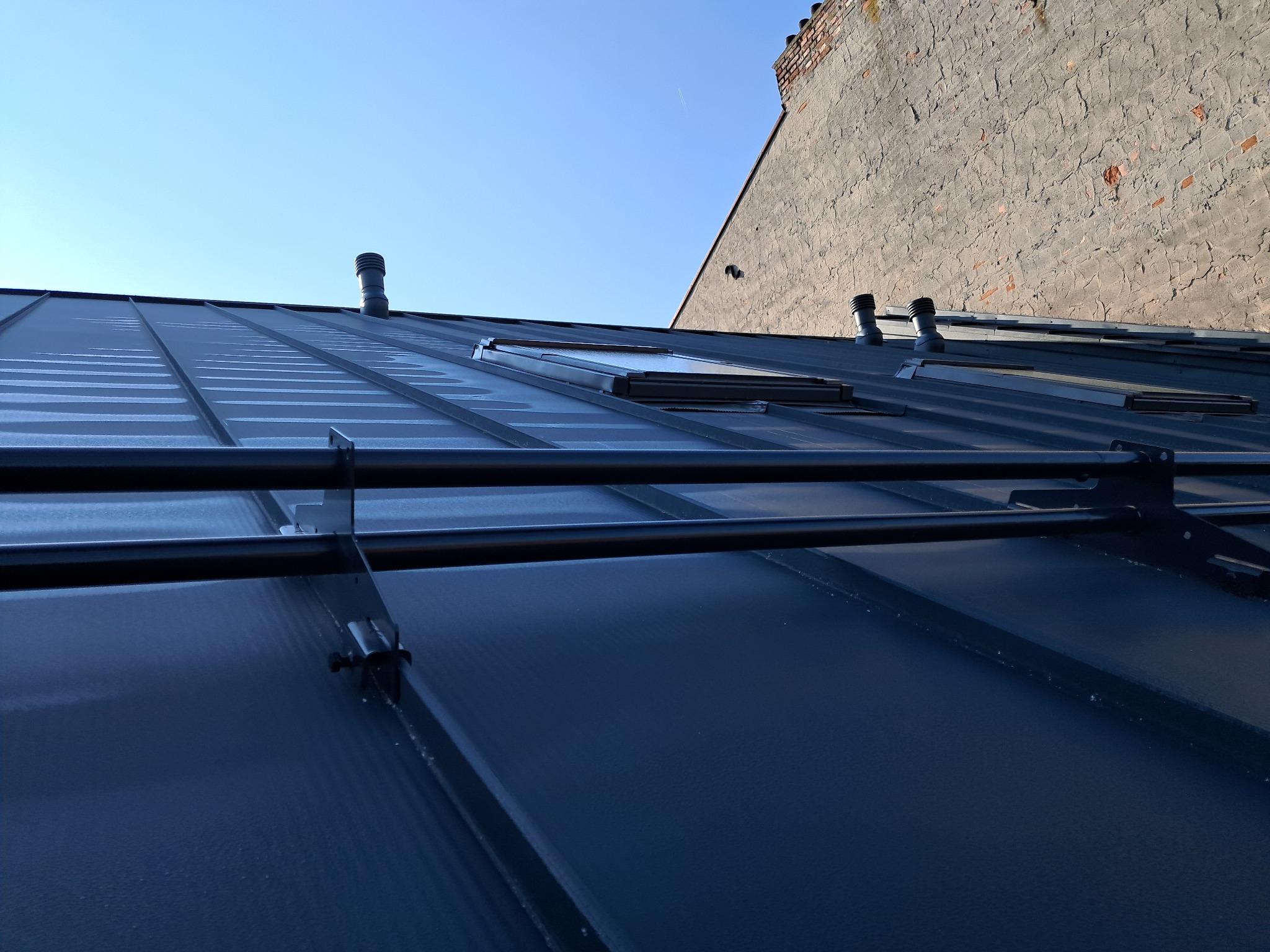 Grafitowy dach z blachy na rąbek stojący z oknami dachowymi i kominkami wentylacyjnymi na tle jasnego nieba i ściany kamienicy.