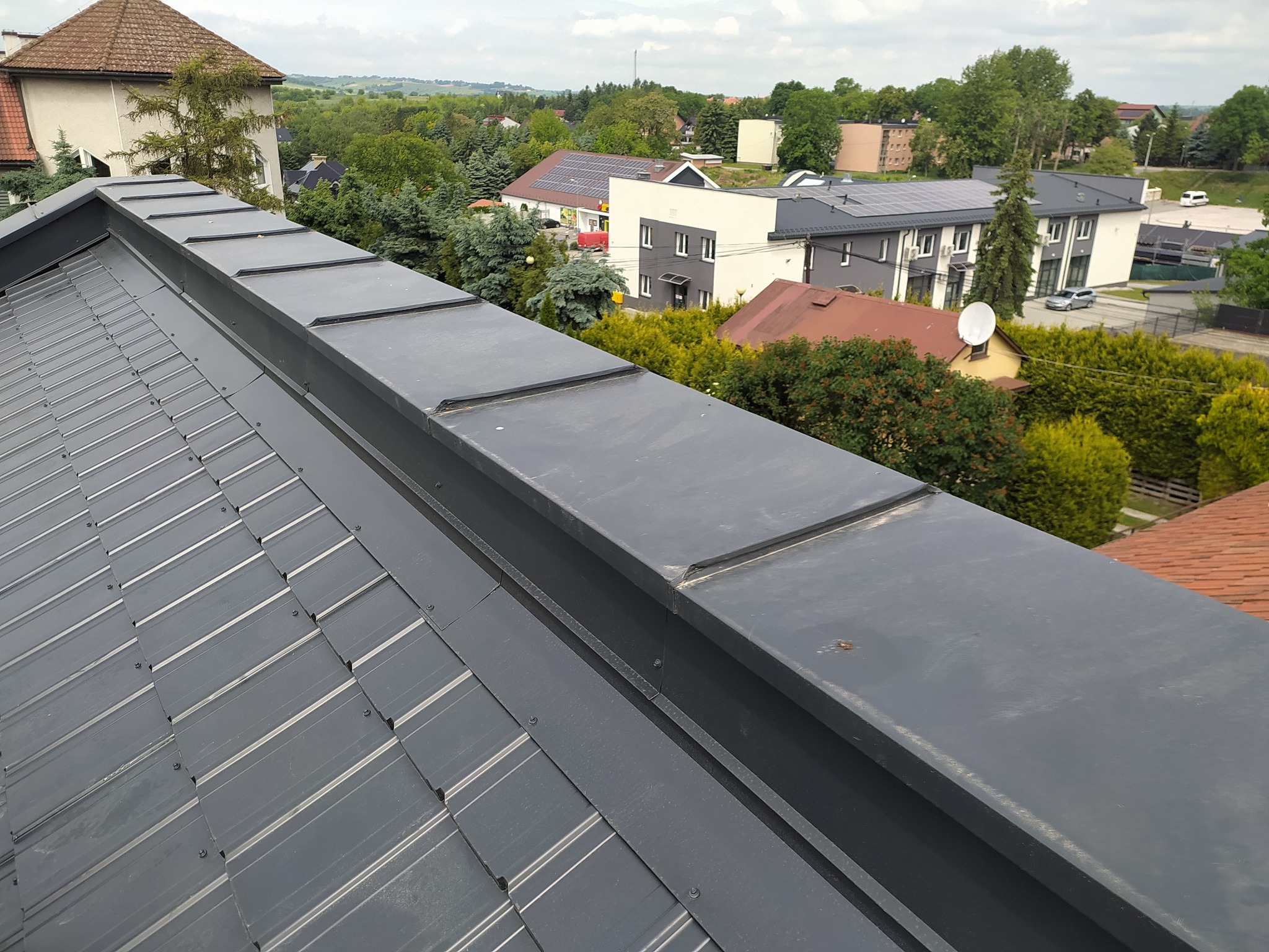 Szary, metalowy dach z widocznymi łączeniami blachy, na tle zieleni i zabudowań, sfotografowany z góry pod lekkim kątem. Widoczny fragment rynny.