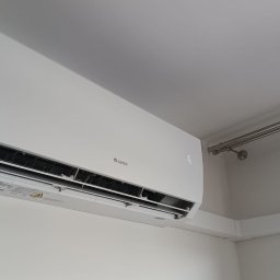 Volta System - Biały klimatyzator marki Gree zamontowany na ścianie, widoczne rury odprowadzające. Minimalistyczny design, jasne wnętrze, kątowy widok z dołu.