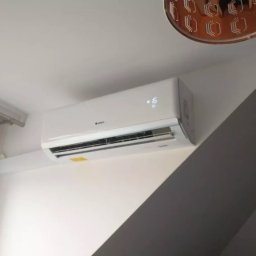 Volta System - Biały klimatyzator ścienny marki Gree zamontowany w rogu pokoju, widoczny fragment okna i lampy sufitowej. Nowoczesny design, czyste wnętrze.