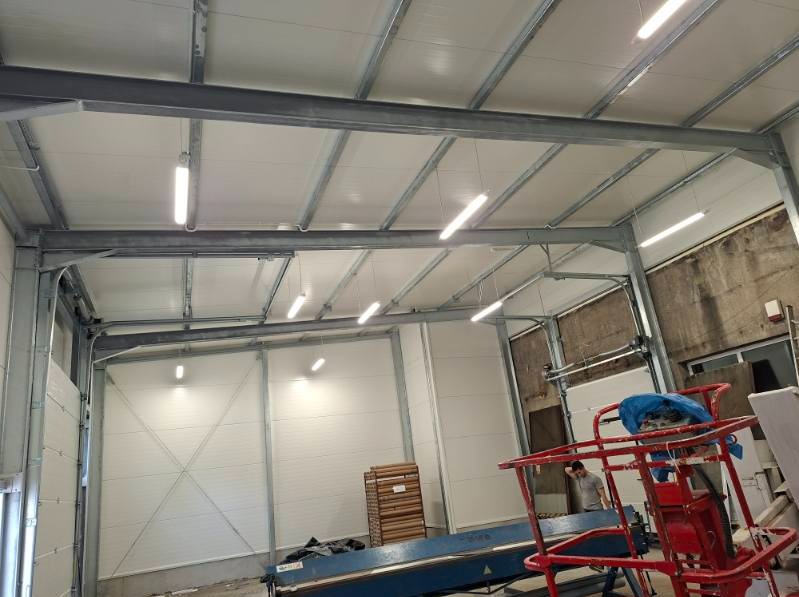 Wnętrze hali z zamontowanymi lampami liniowymi pod sufitem, widoczne metalowe konstrukcje i białe ściany. Na podłodze maszyny i materiały budowlane.