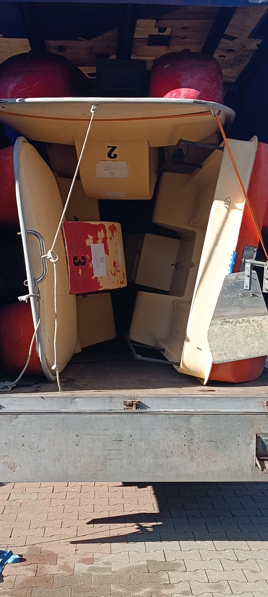 Załadunek łodzi i boi do transportu na przyczepie. Widoczne numery '2' i '3' na elementach ładunku. Przyczepa na kostce brukowej.