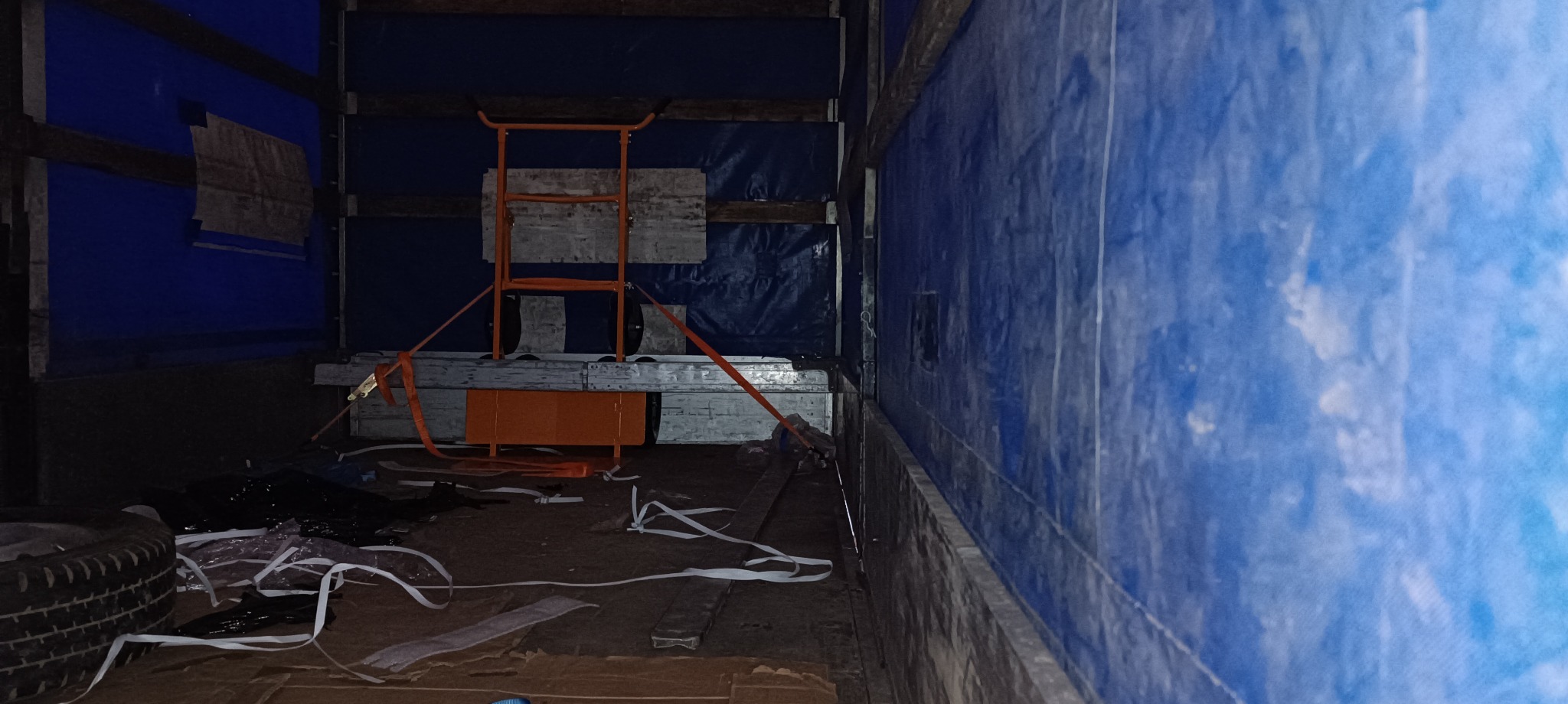 Wnętrze przestrzeni ładunkowej busa z zabezpieczonymi elementami, w tym pomarańczowym wózkiem transportowym i oponą. Widoczne pasy mocujące i plandeka.