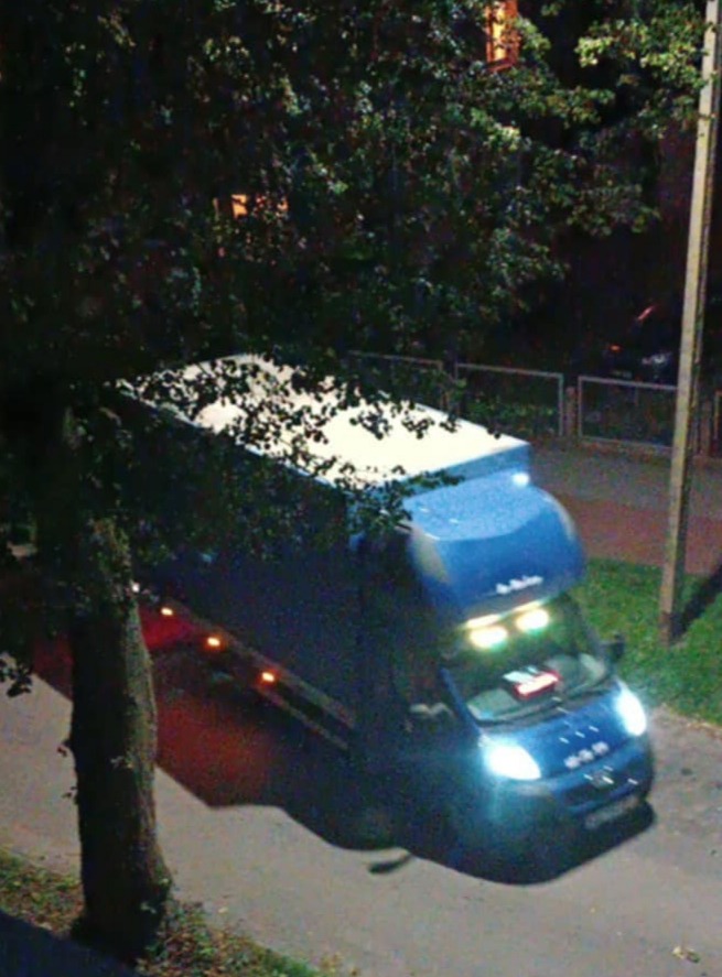 Niebieski bus z włączonymi światłami, widziany nocą z góry, częściowo zasłonięty przez drzewo. Biały dach i ciemne otoczenie.