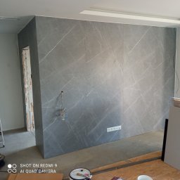 Monolit - Ściana wykończona panelami imitującymi marmur z widocznymi gniazdkami, przewody elektryczne i fragmentem ościeżnicy w trakcie prac wykończeniowych.