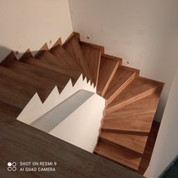 Monolit - Wysoki kąt na drewniane schody spiralne z białymi ścianami. Widoczne niedoskonałości na ścianie, sugerujące etap wykończeniowy. Ciepłe oświetlenie.