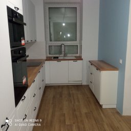 Monolit - Nowoczesna kuchnia z białymi szafkami, drewnianym blatem i parkietem, oświetlona naturalnym światłem wpadającym przez okno z żaluzjami.