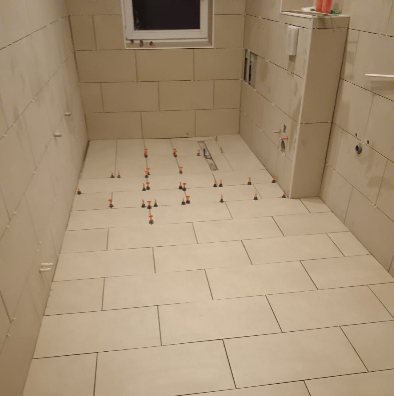 Beżowe płytki podłogowe w łazience, z systemem poziomowania płytek. Widoczny odpływ liniowy, ściany częściowo wyłożone płytkami. Ujęcie z góry, ukośne.