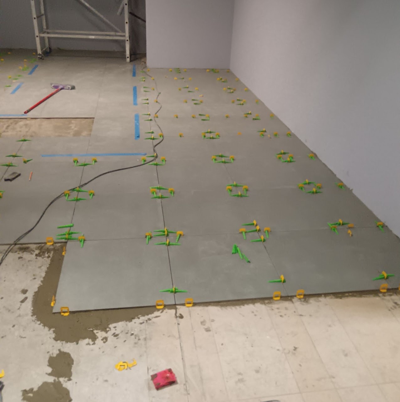 Układanie szarych płytek podłogowych z użyciem poziomujących klipsów i klinów. Widoczna zaprawa i fragment rusztowania.