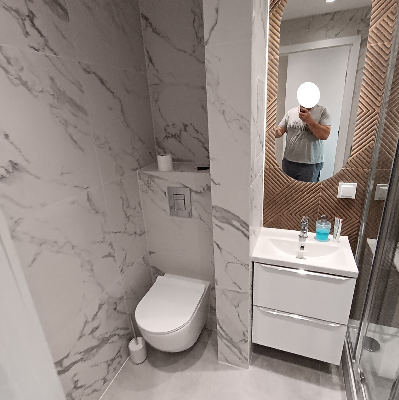 Nowoczesna łazienka z marmurowymi płytkami, owalnym lustrem z dekoracyjną ramą i białą szafką pod umywalką. Widoczny fragment kabiny prysznicowej.