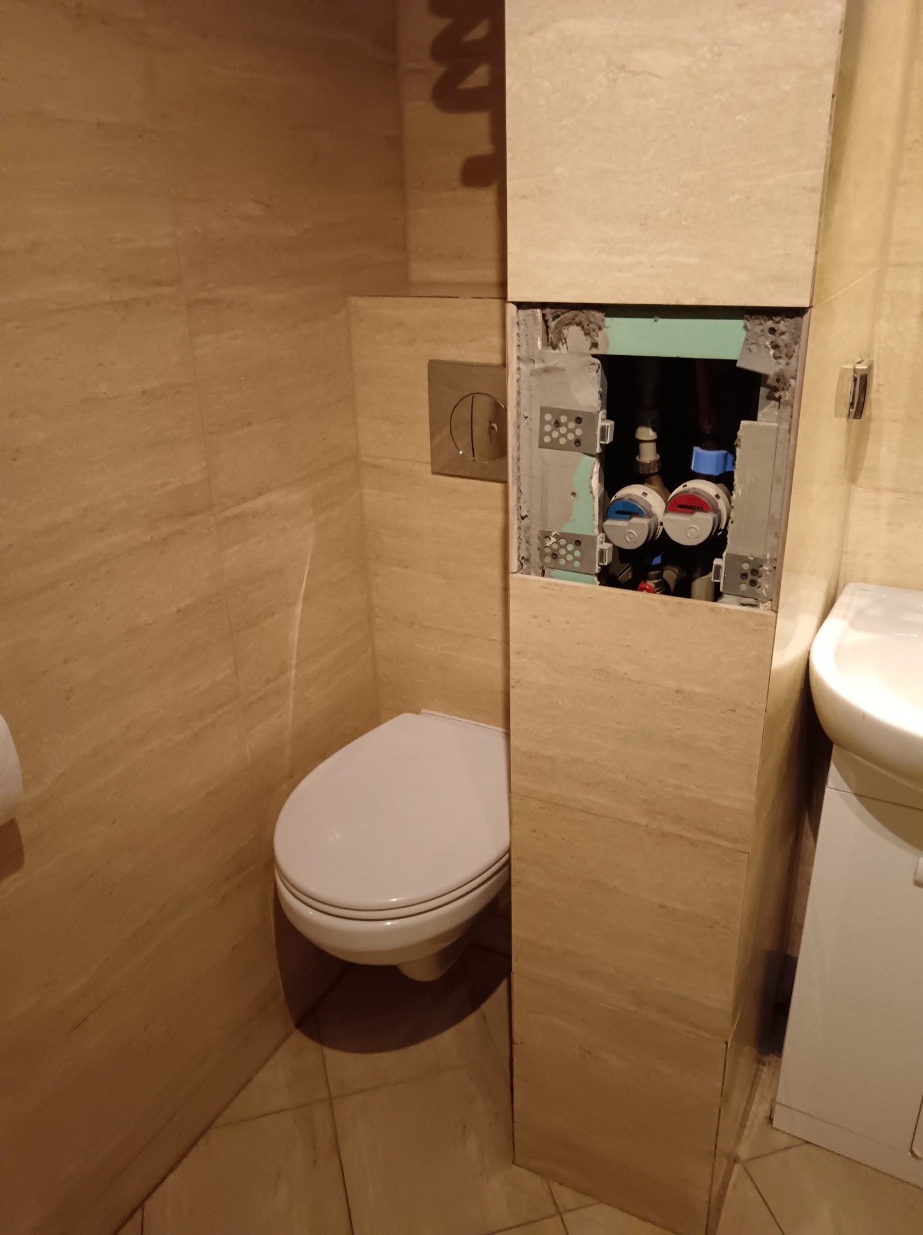 Łazienka w trakcie remontu: widoczny sedes i zabudowa z odkrytymi zaworami wody, płytki imitujące marmur na ścianach i podłodze, umywalka z szafką obok.