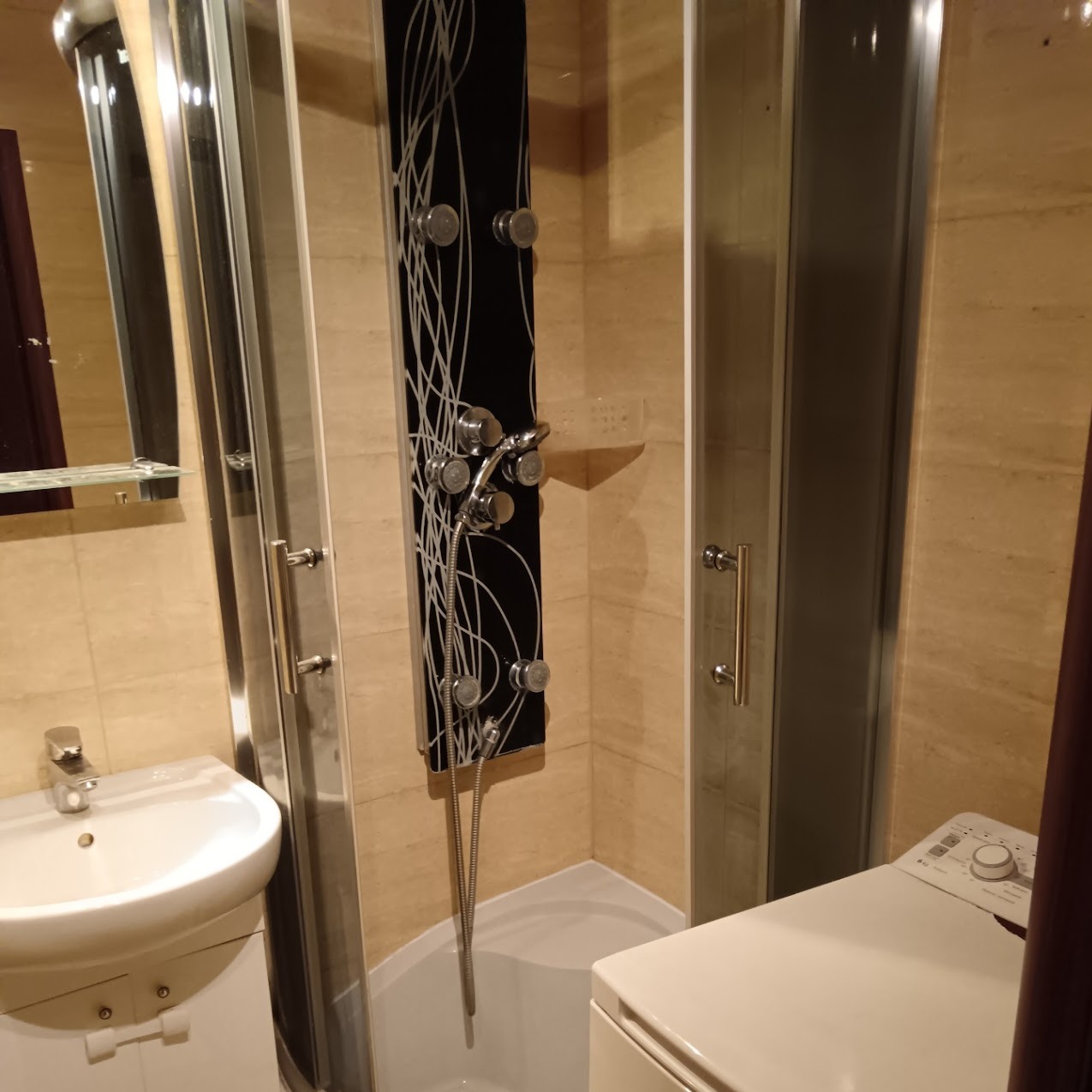 Kompaktowa łazienka z kabiną prysznicową z hydromasażem, umywalką i pralką. Beżowe płytki na ścianach. Elementy czarne i srebrne.