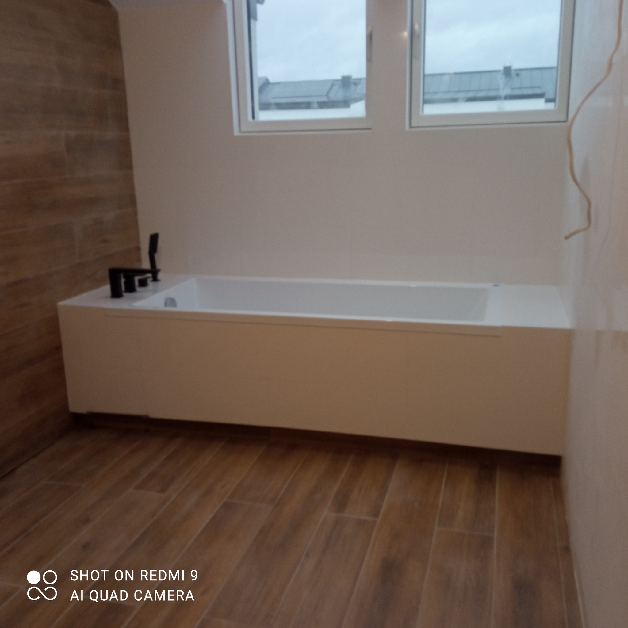 Wykończona łazienka z drewnopodobnymi płytkami na podłodze i ścianie, białą wanną z czarną armaturą. Dwa okna w tle wpuszczają naturalne światło.