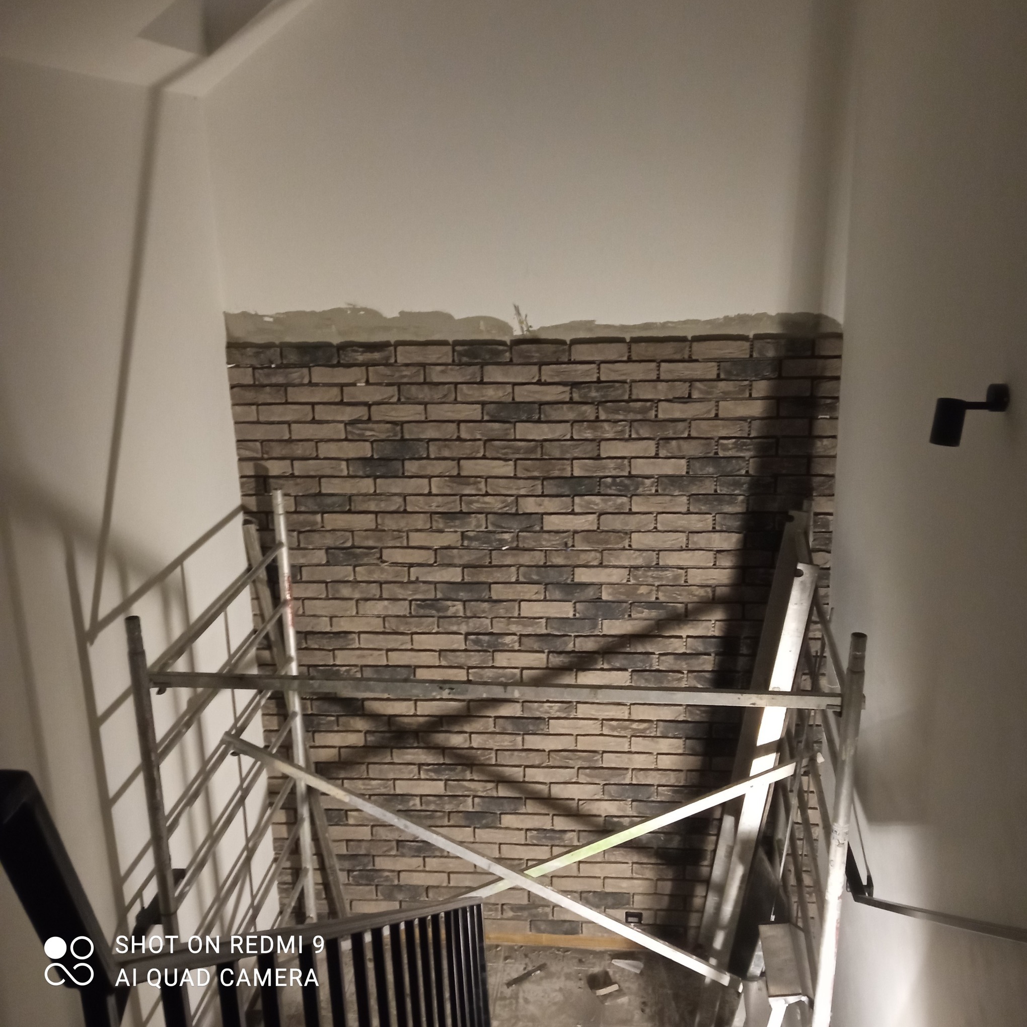 Ściana wyłożona cegłą klinkierową na klatce schodowej, widoczny fragment rusztowania. Praca w toku, oświetlenie punktowe.