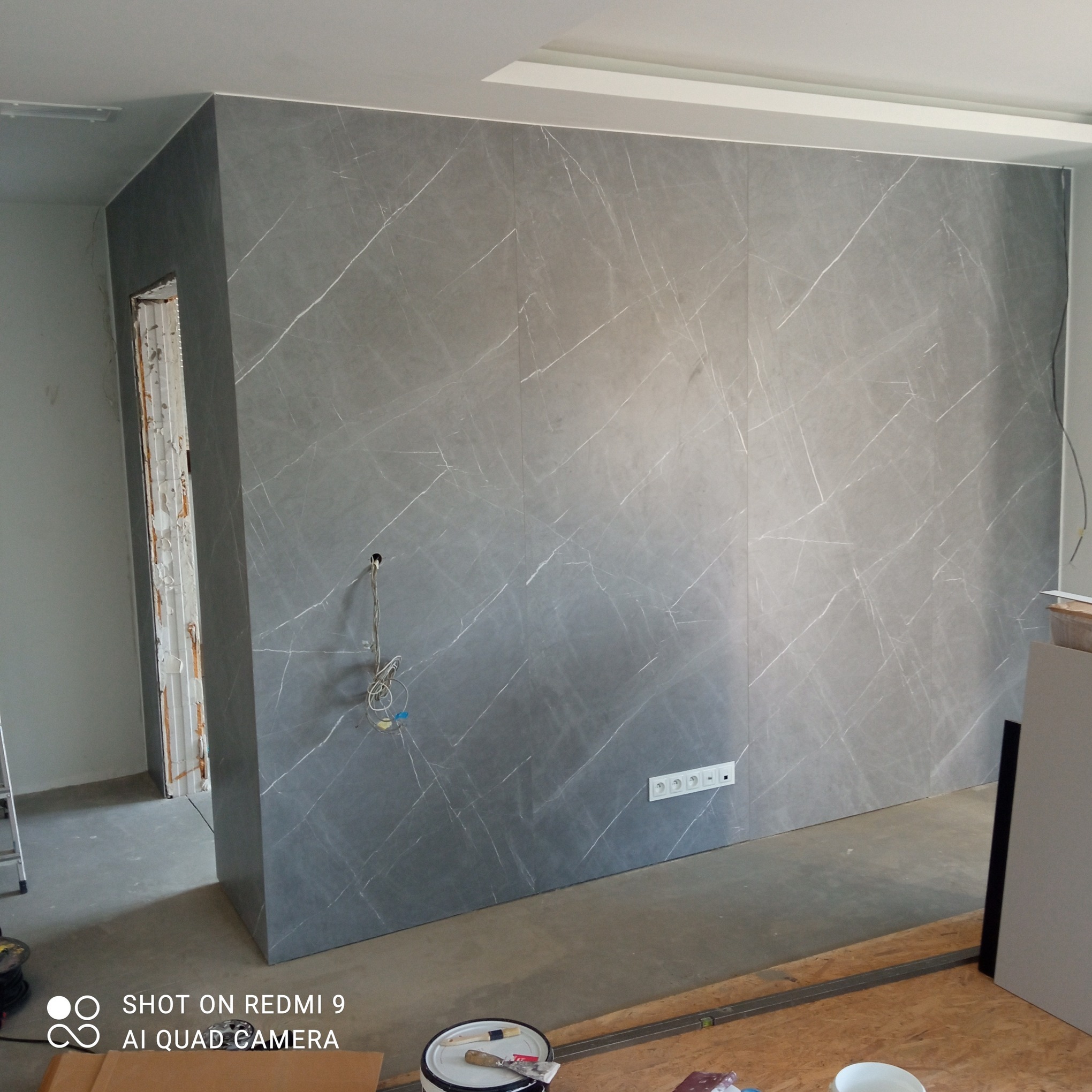 Ściana wykończona panelami imitującymi marmur z widocznymi gniazdkami, przewody elektryczne i fragmentem ościeżnicy w trakcie prac wykończeniowych.