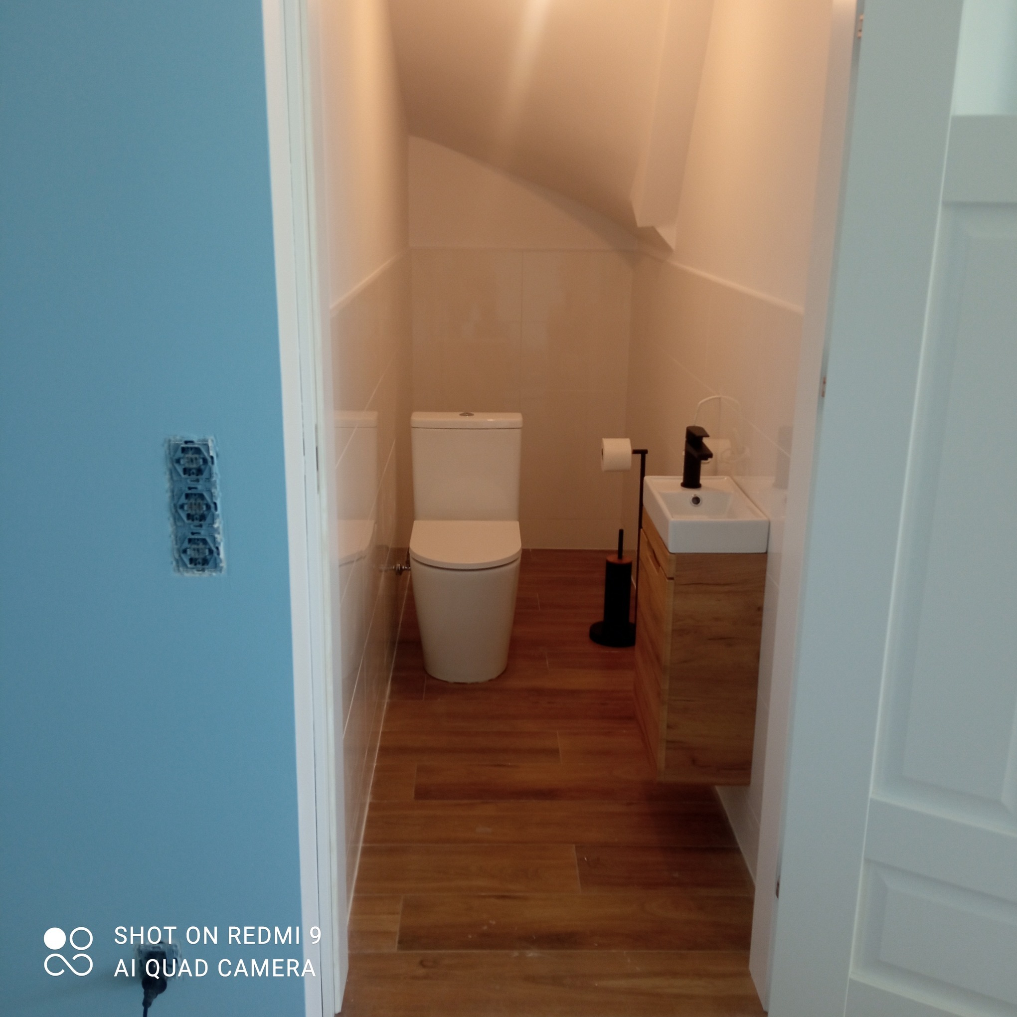 Nowoczesna łazienka na poddaszu z drewnopodobną podłogą, białą ceramiką i umywalką z szafką. Na ścianie widoczna instalacja elektryczna bez wykończenia.