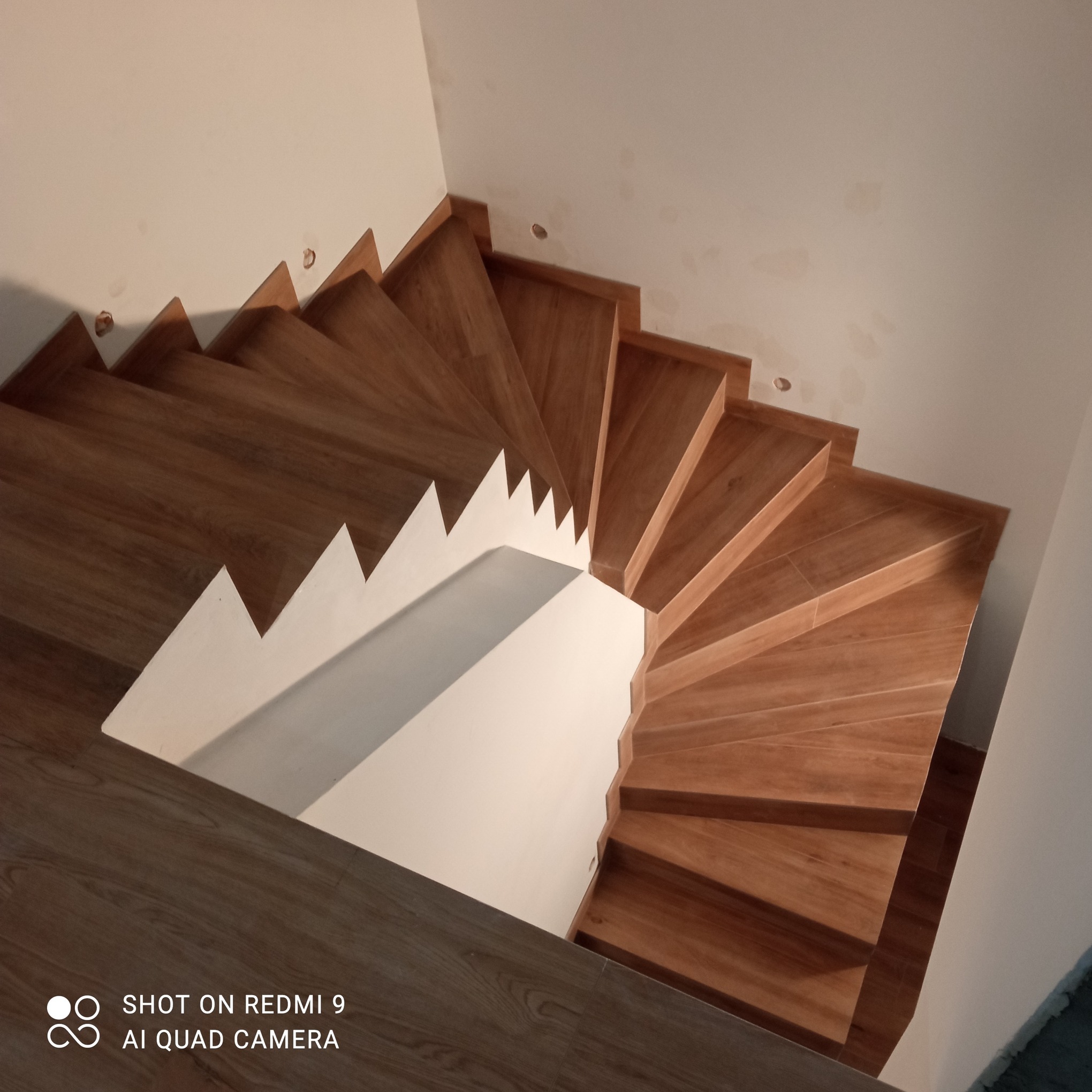 Wysoki kąt na drewniane schody spiralne z białymi ścianami. Widoczne niedoskonałości na ścianie, sugerujące etap wykończeniowy. Ciepłe oświetlenie.