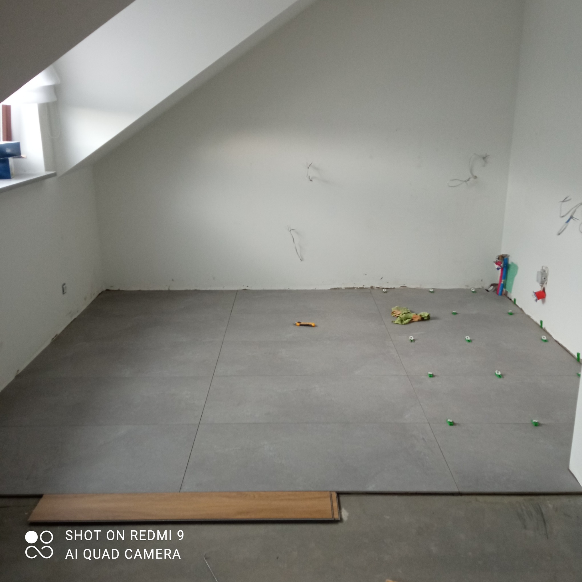 Szare płytki podłogowe ułożone w pomieszczeniu ze skośnym sufitem, widoczne narzędzia glazurnicze i wystające kable elektryczne.