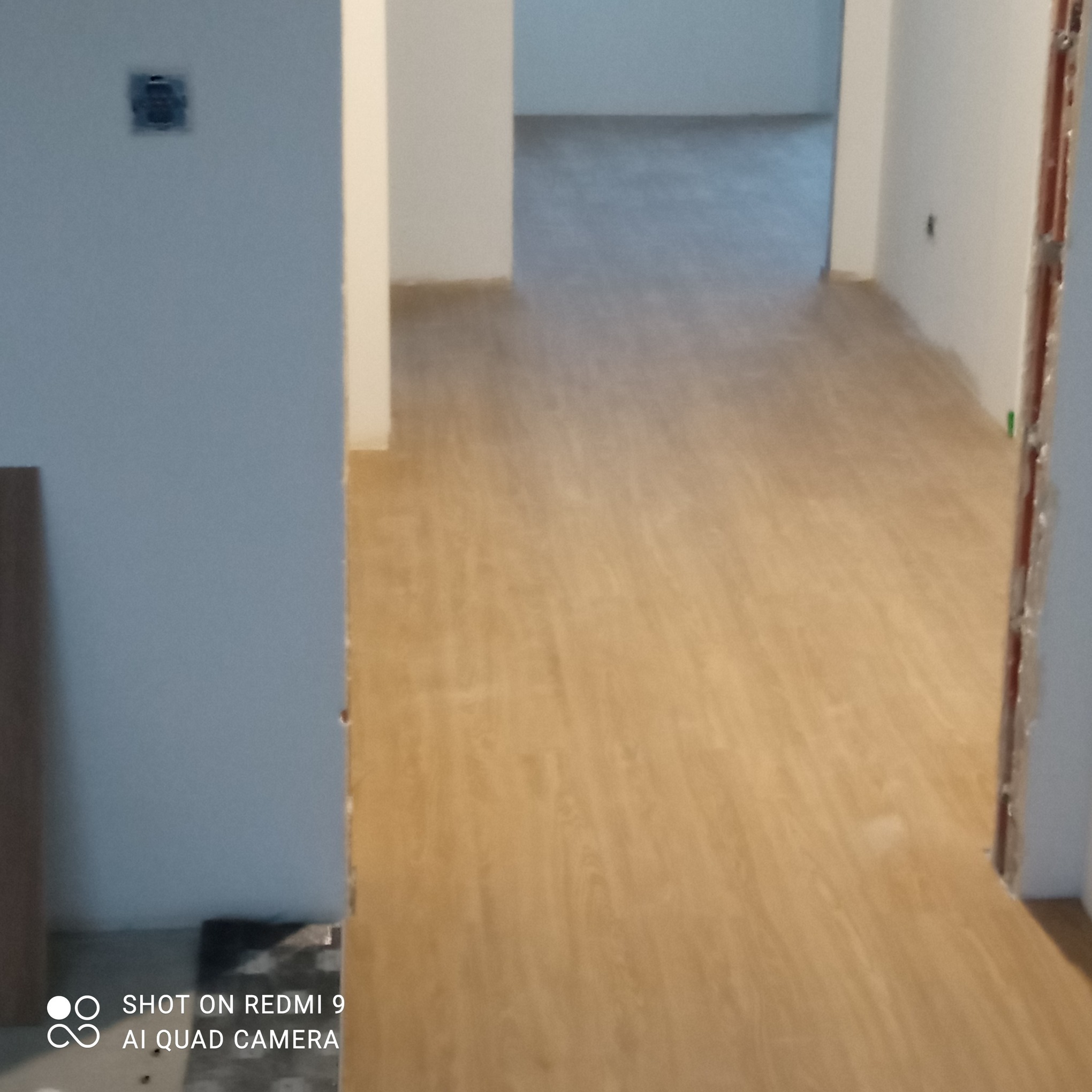 Jasne panele podłogowe z widocznym wzorem drewna w nowym mieszkaniu. Na ścianie puszka elektryczna bez osprzętu. Widok na kolejne pomieszczenia.