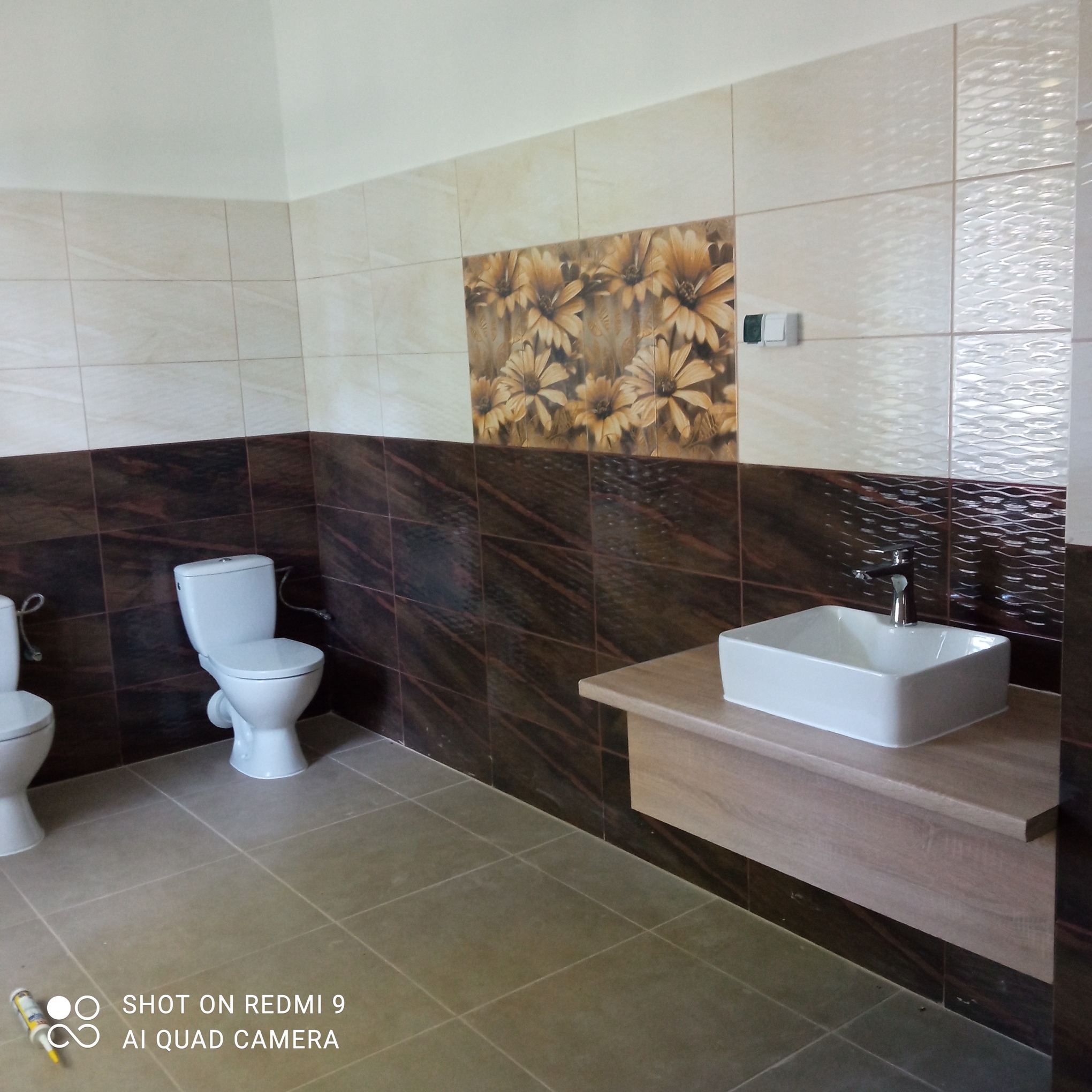 Wykończona łazienka z beżowo-brązowymi płytkami, umywalką na szafce z imitacją drewna i toaletami. Na podłodze szare płytki. Widoczny silikon sanitarny.