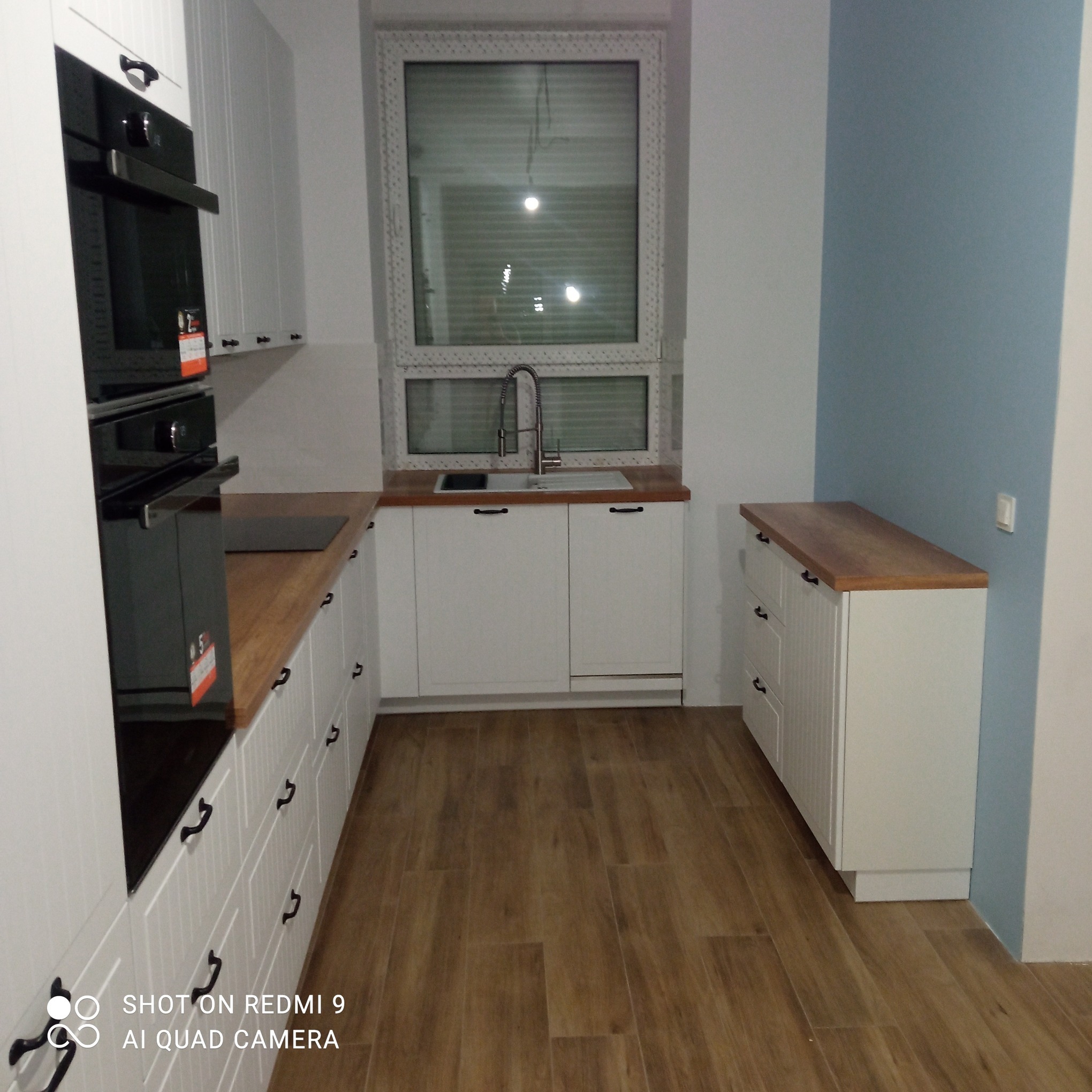 Nowoczesna kuchnia z białymi szafkami, drewnianym blatem i parkietem, oświetlona naturalnym światłem wpadającym przez okno z żaluzjami.