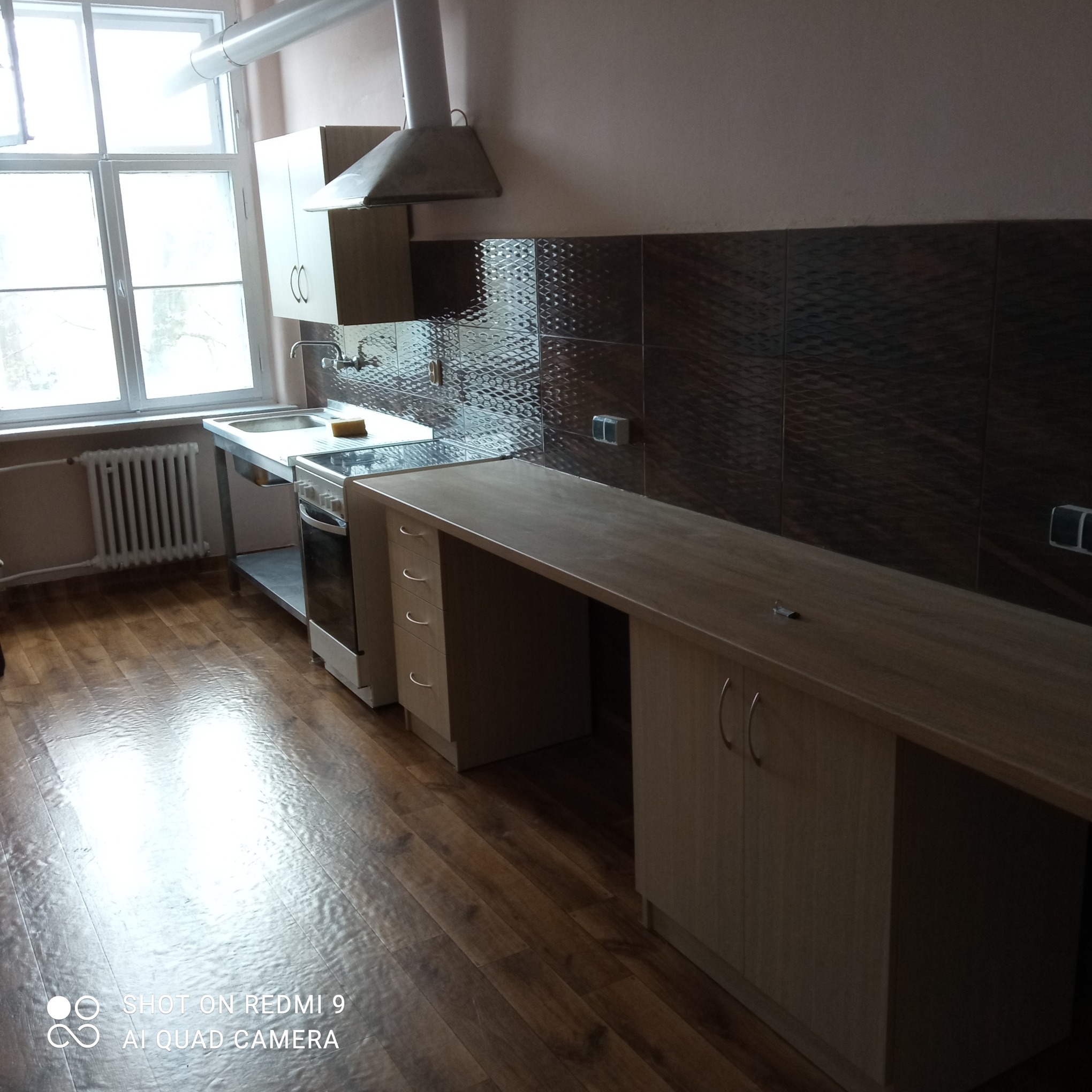 Ukończona kuchnia z drewnianą podłogą, zabudową meblową i błyszczącymi, ciemnymi płytkami na ścianie. Widok na okno i grzejnik. Metalowy zlew i kuchenka.