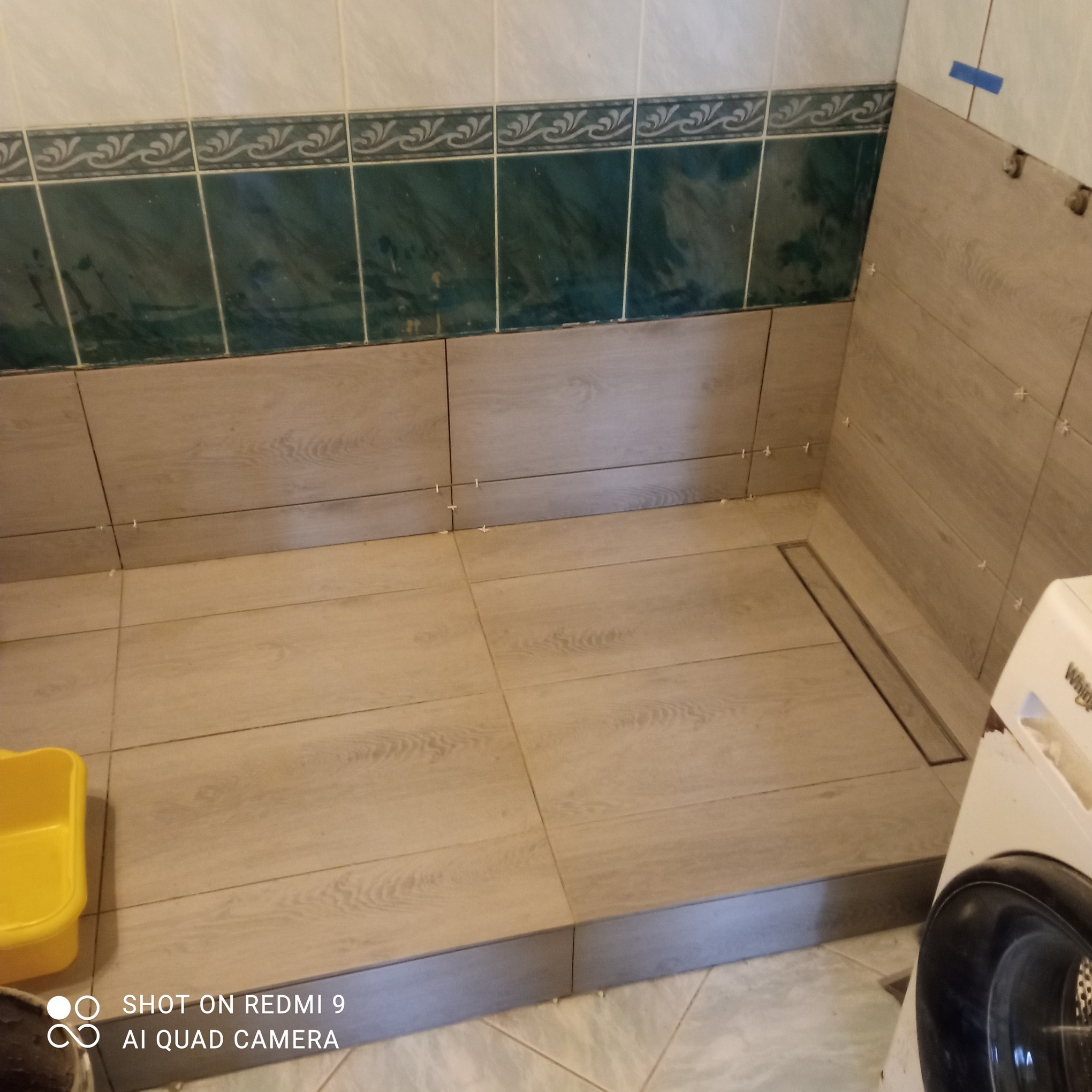 Łazienka w trakcie remontu: ułożone płytki podłogowe imitujące drewno, widoczny odpływ liniowy, częściowo położone płytki ścienne z dekoracyjnym paskiem.