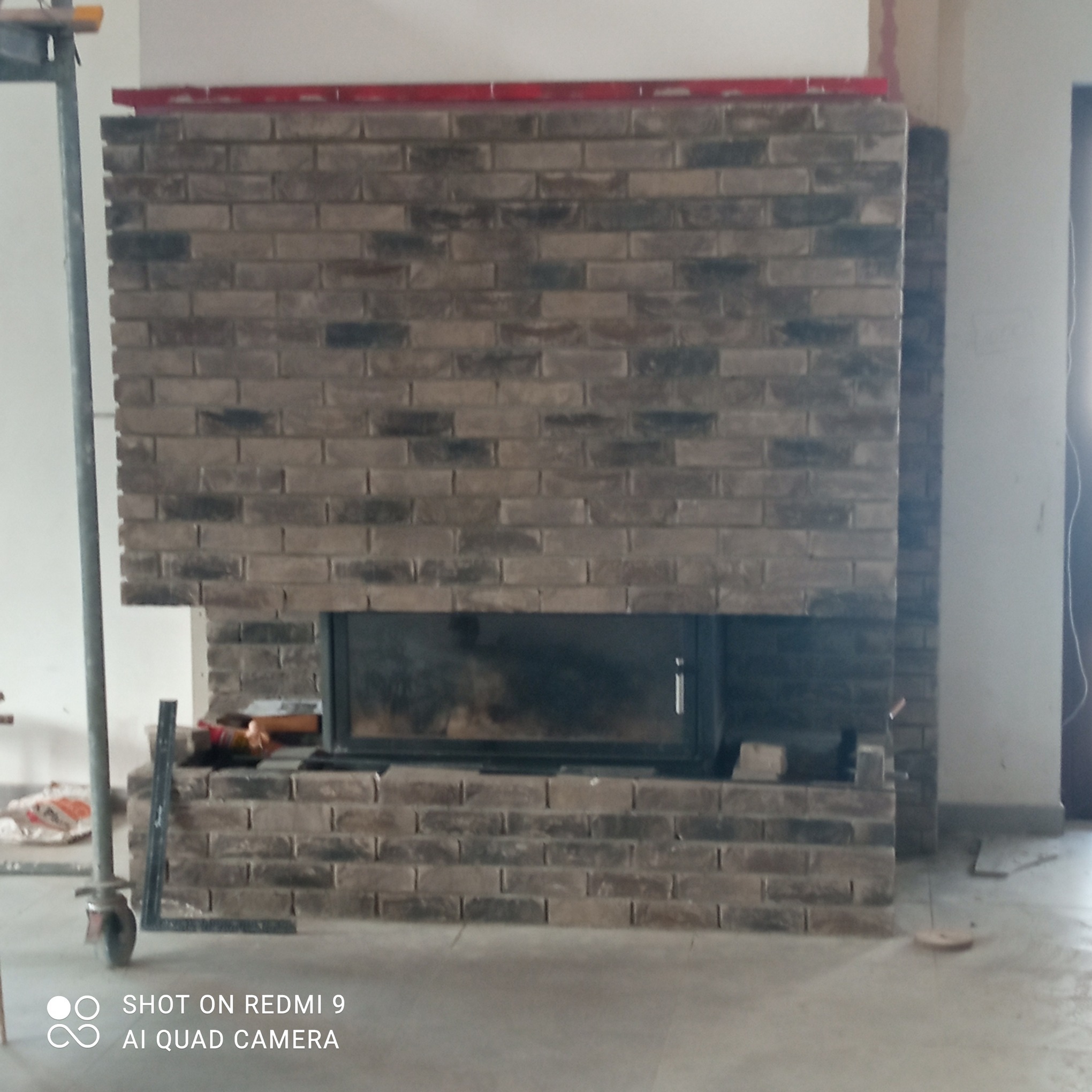 Wnętrze z kominkiem obłożonym cegłą klinkierową w trakcie montażu. Widoczny poziomica, rusztowanie i narzędzia. Surowe wykończenie, etap prac wykończeniowych.