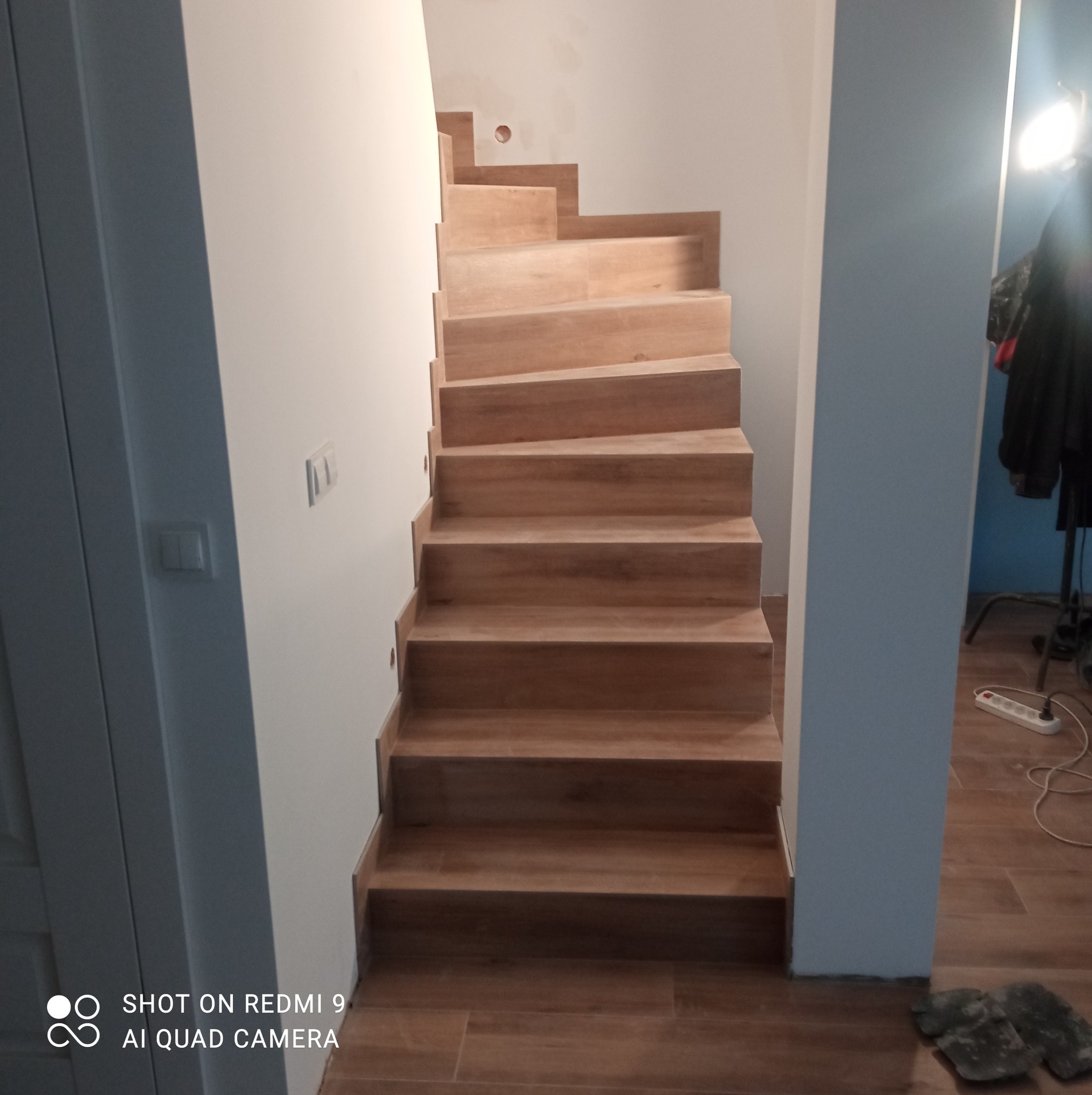 Wykończone schody drewniane z widocznymi stopniami i podstopnicami, prowadzące do góry. Na podłodze płytki imitujące drewno. W tle widoczny fragment ściany i lampa.