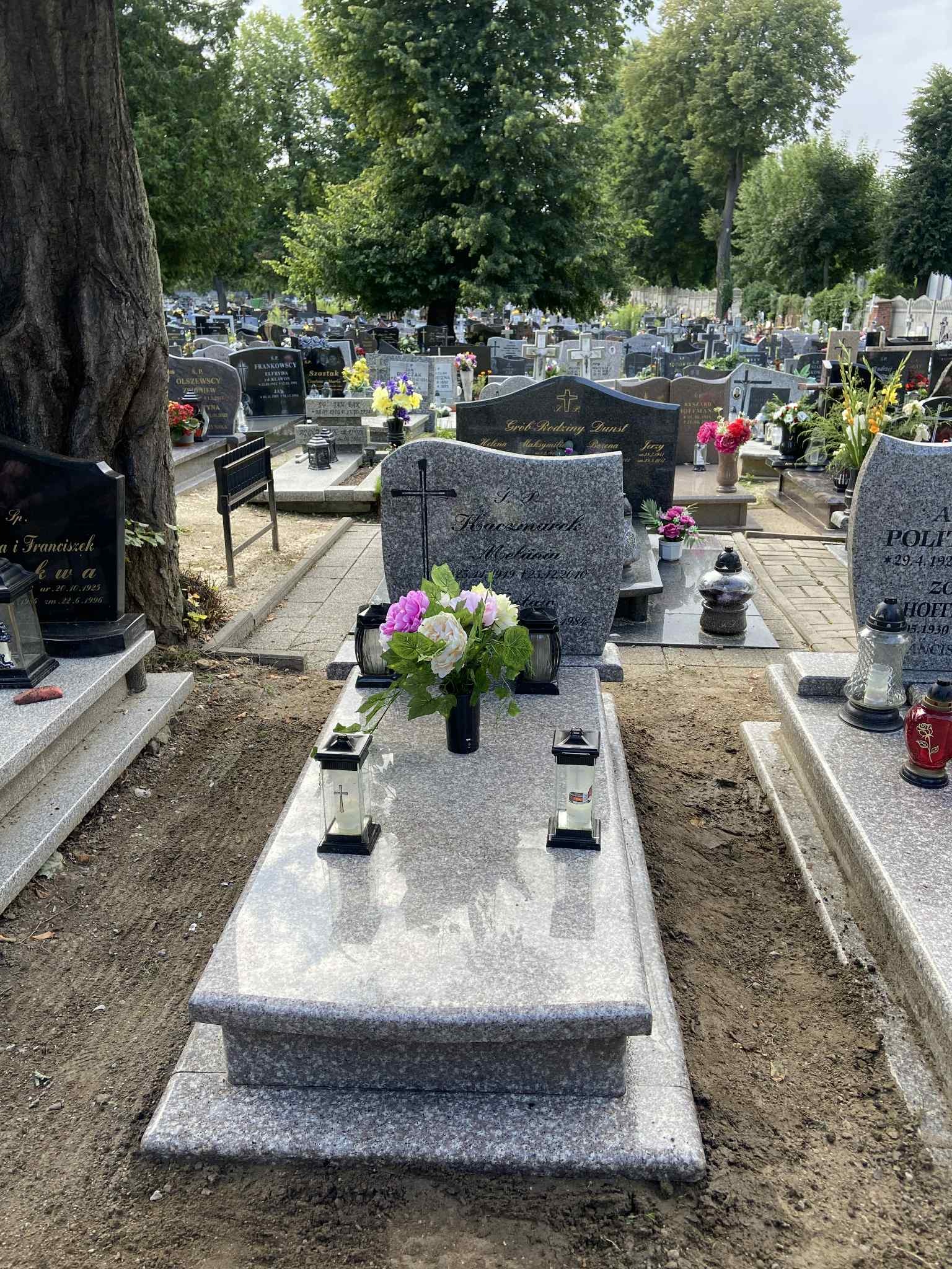 Jasnoszary nagrobek z granitu na cmentarzu, ozdobiony kwiatami i lampionami. Widok na inne groby i drzewa w tle.