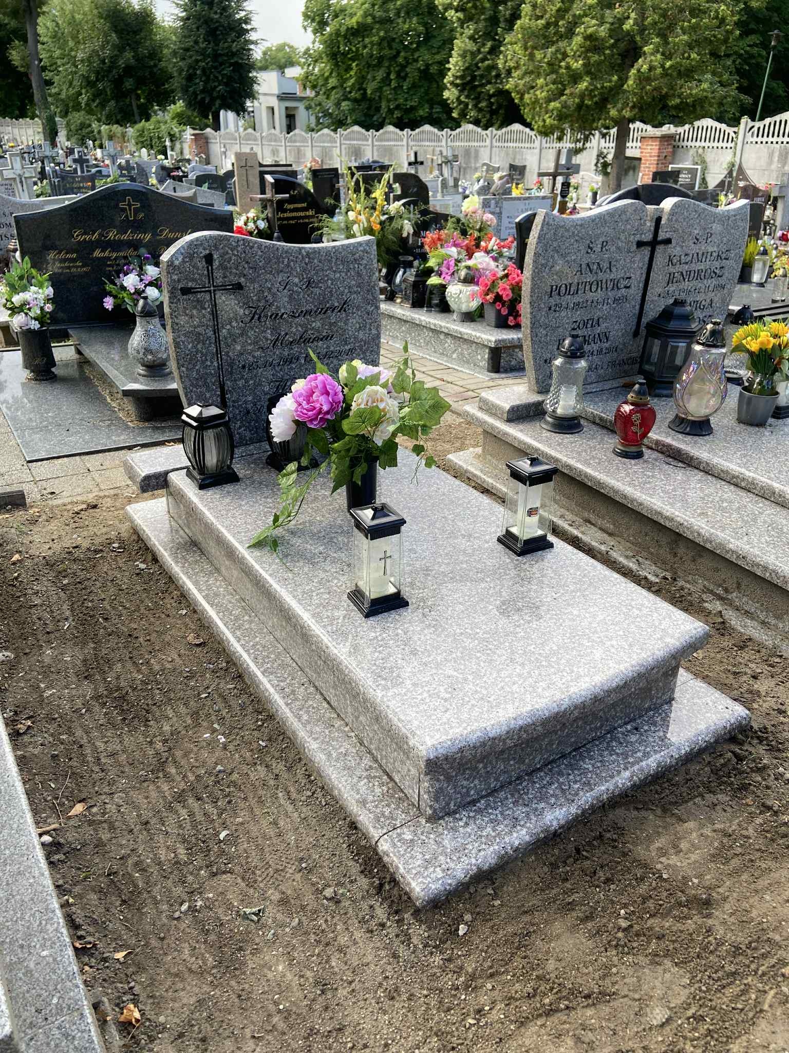 Jasnoszary, dwupoziomowy pomnik z granitu na cmentarzu, ozdobiony kwiatami i lampionami. Widoczne nagrobki w tle, zieleń drzew i fragment ogrodzenia.