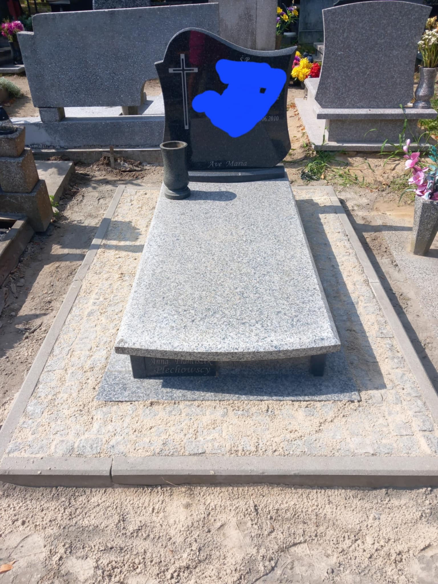 Jasnoszary nagrobek z ciemną tablicą i wazonem, otoczony piaskiem i kostką. Widok z przodu, nagrobek na cmentarzu.