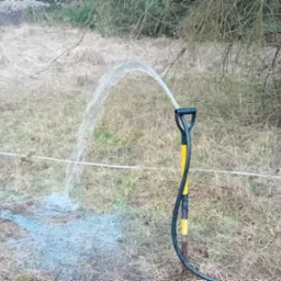 Test wydajności pompy studni głębinowej: woda tryska z węża na tle trawy i drzewa. Czarny wąż z żółtymi detalami wbity w ziemię.