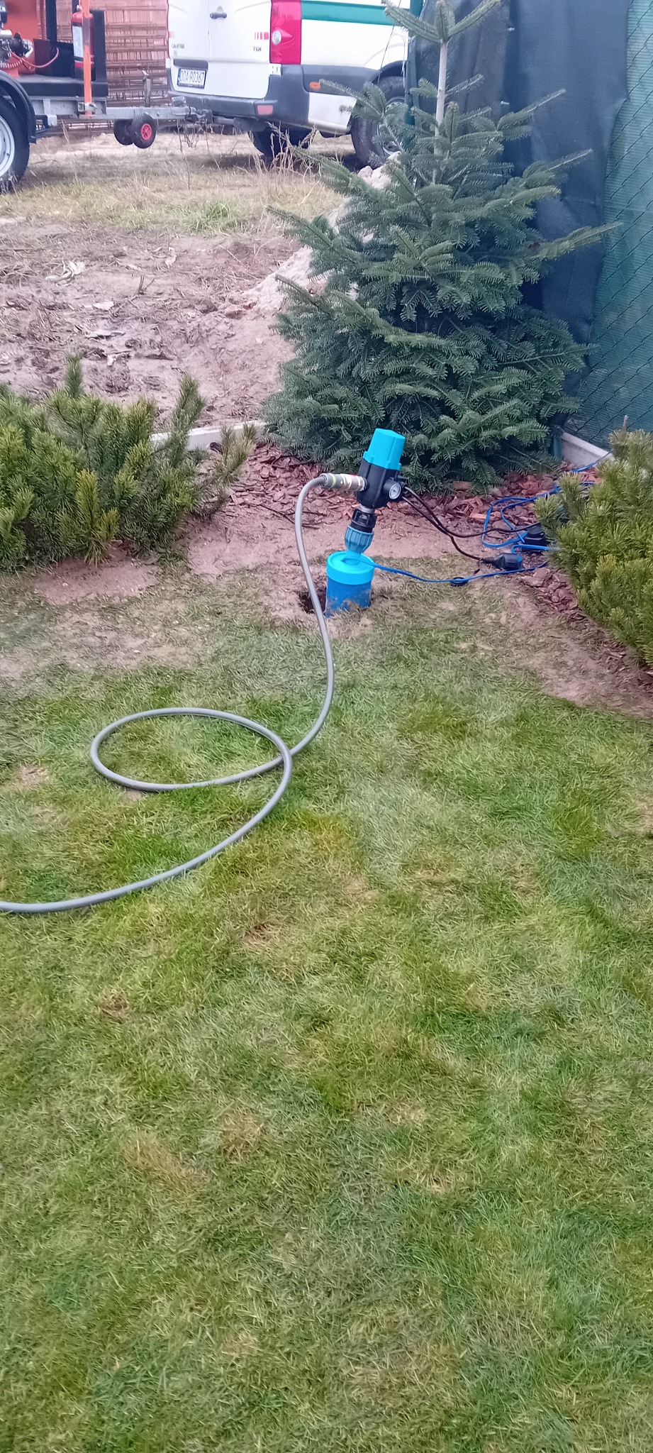 Niebieska obudowa studni abisyńskiej z podłączonymi przewodami na trawniku, obok iglaków i zaparkowanego pojazdu z przyczepką. Widok z góry.