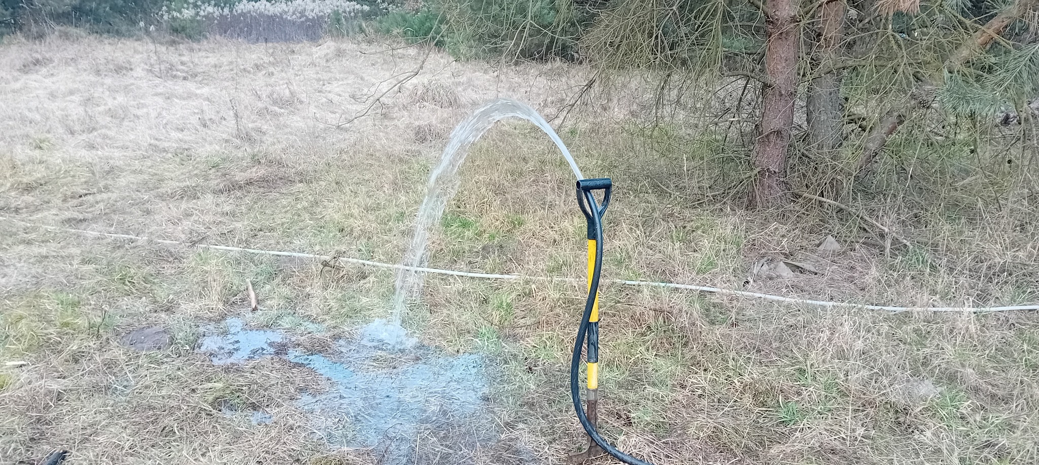 Test wydajności pompy studni głębinowej: woda tryska z węża na tle trawy i drzewa. Czarny wąż z żółtymi detalami wbity w ziemię.
