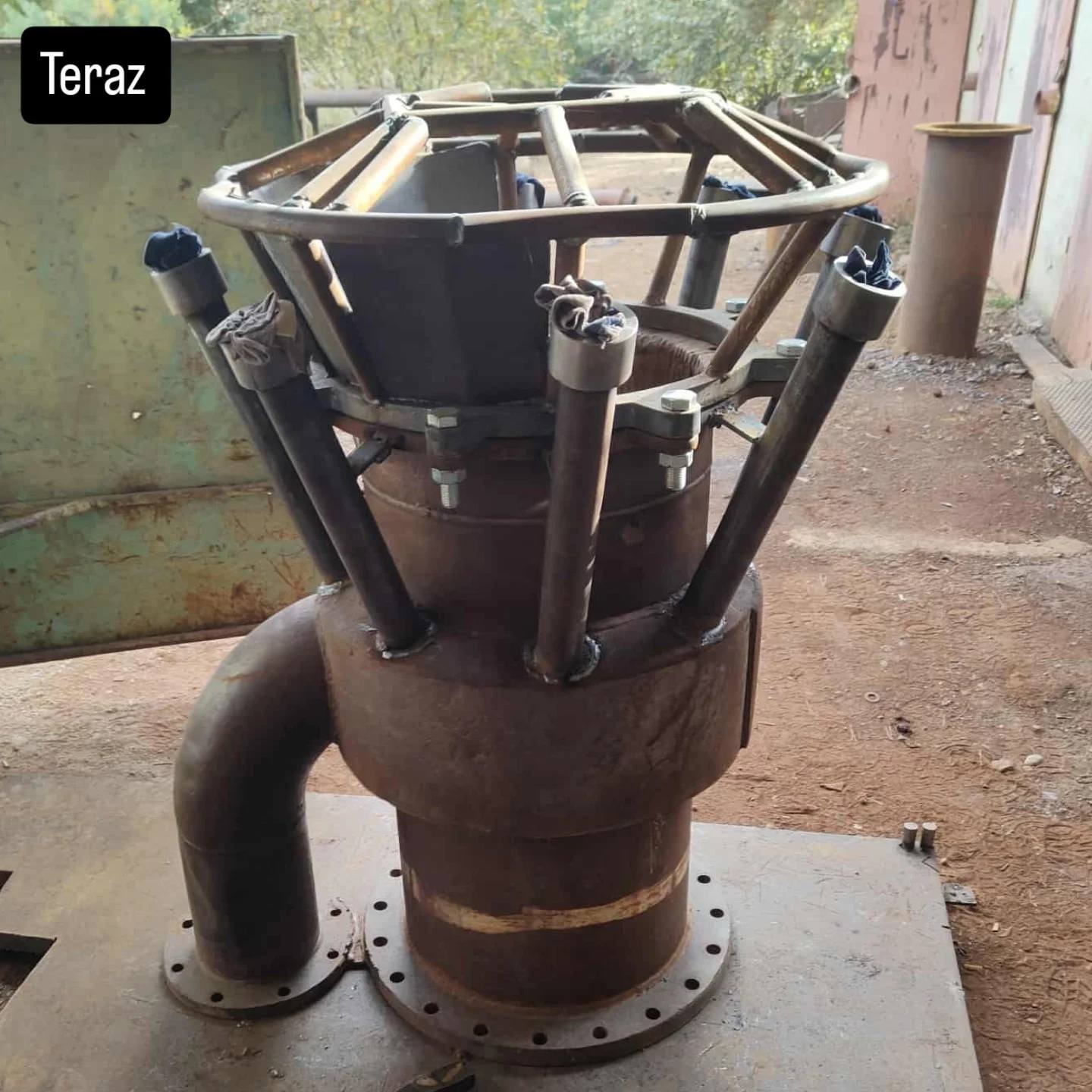 Surowa, spawana konstrukcja metalowa z rur i profili, z widocznymi spawami i elementami montażowymi, umieszczona na metalowej płycie. Nietypowy kształt.