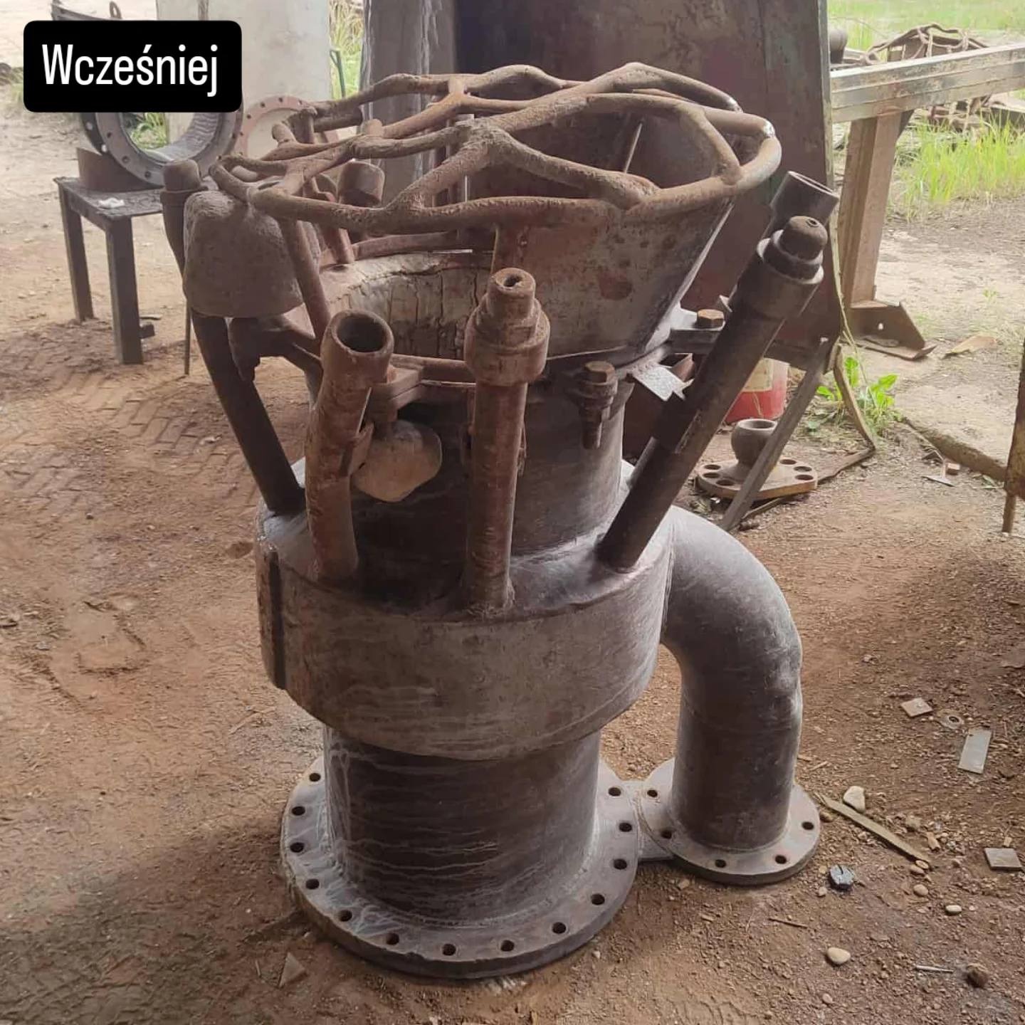 Zardzewiała, metalowa konstrukcja przemysłowa z zaworem kołowym i rurami, stojąca na ziemi. Widoczny napis 'Wcześniej'.