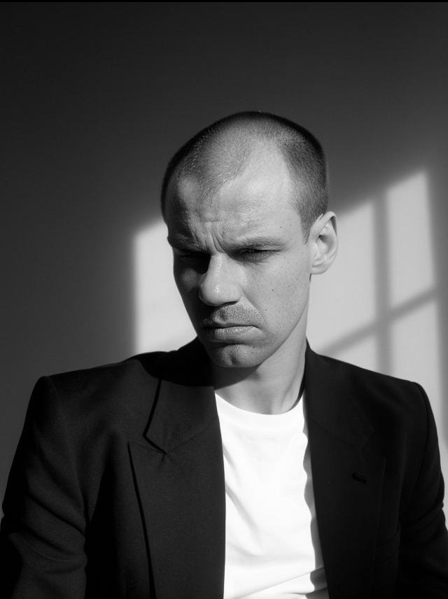 Czarno-biały portret mężczyzny w marynarce, z poważnym wyrazem twarzy, oświetlonego promieniami słońca padającymi przez okno.