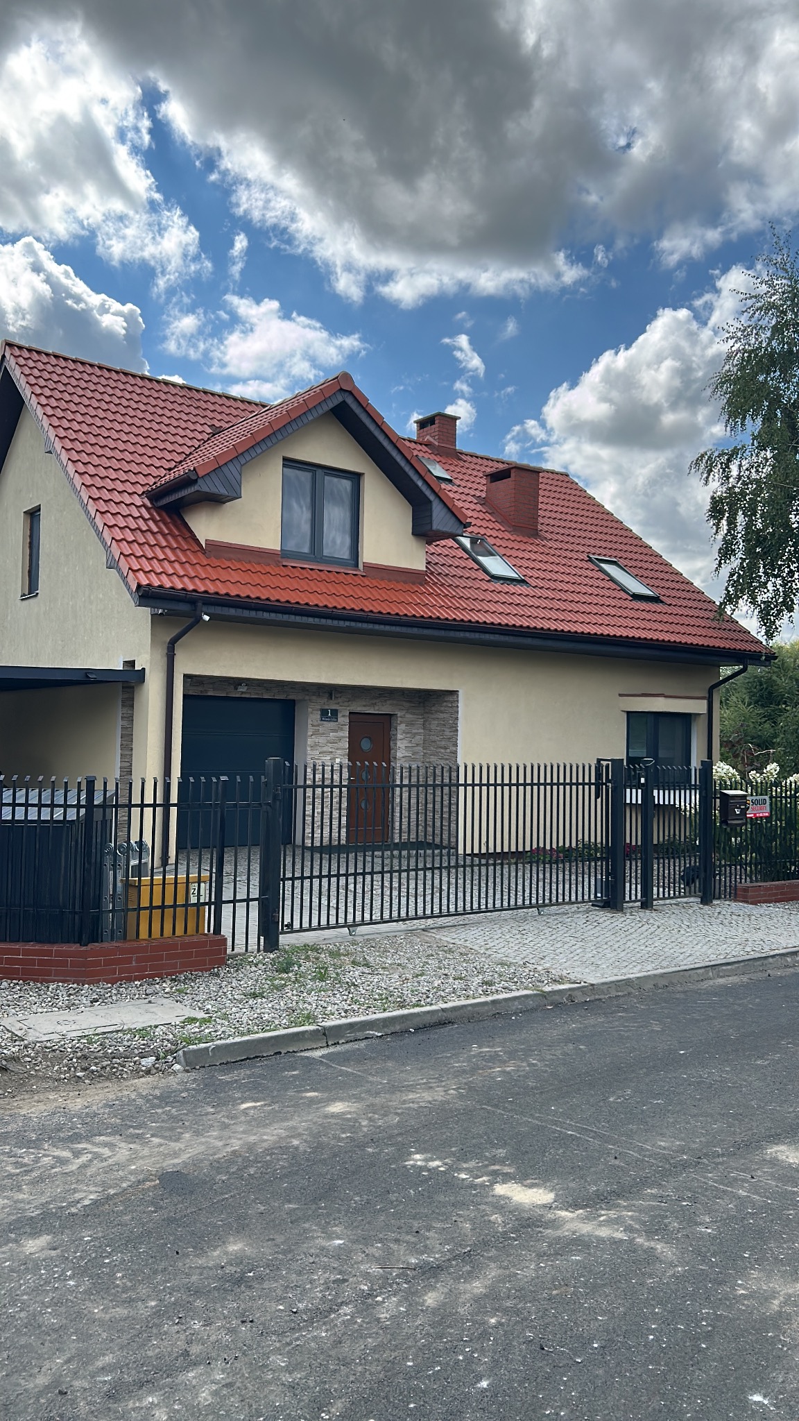 Dom jednorodzinny z czerwonym dachem i beżową elewacją, widoczny z ulicy za czarnym metalowym ogrodzeniem. Na dachu okna połaciowe i komin.