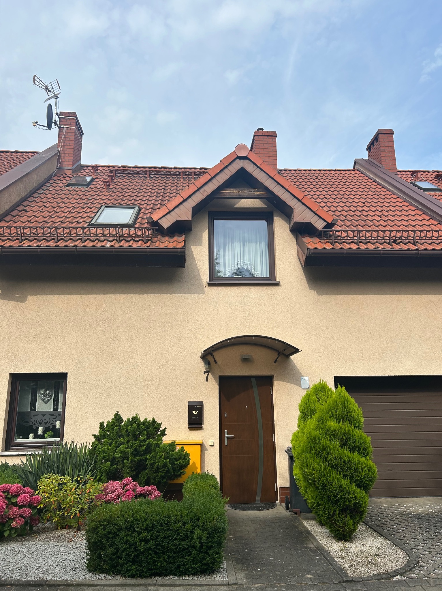 Dwukondygnacyjny dom z czerwoną dachówką i beżową elewacją. Widoczne okna, drzwi wejściowe, garaż i zadbany ogródek z krzewami i kwiatami. Antena satelitarna na dachu.