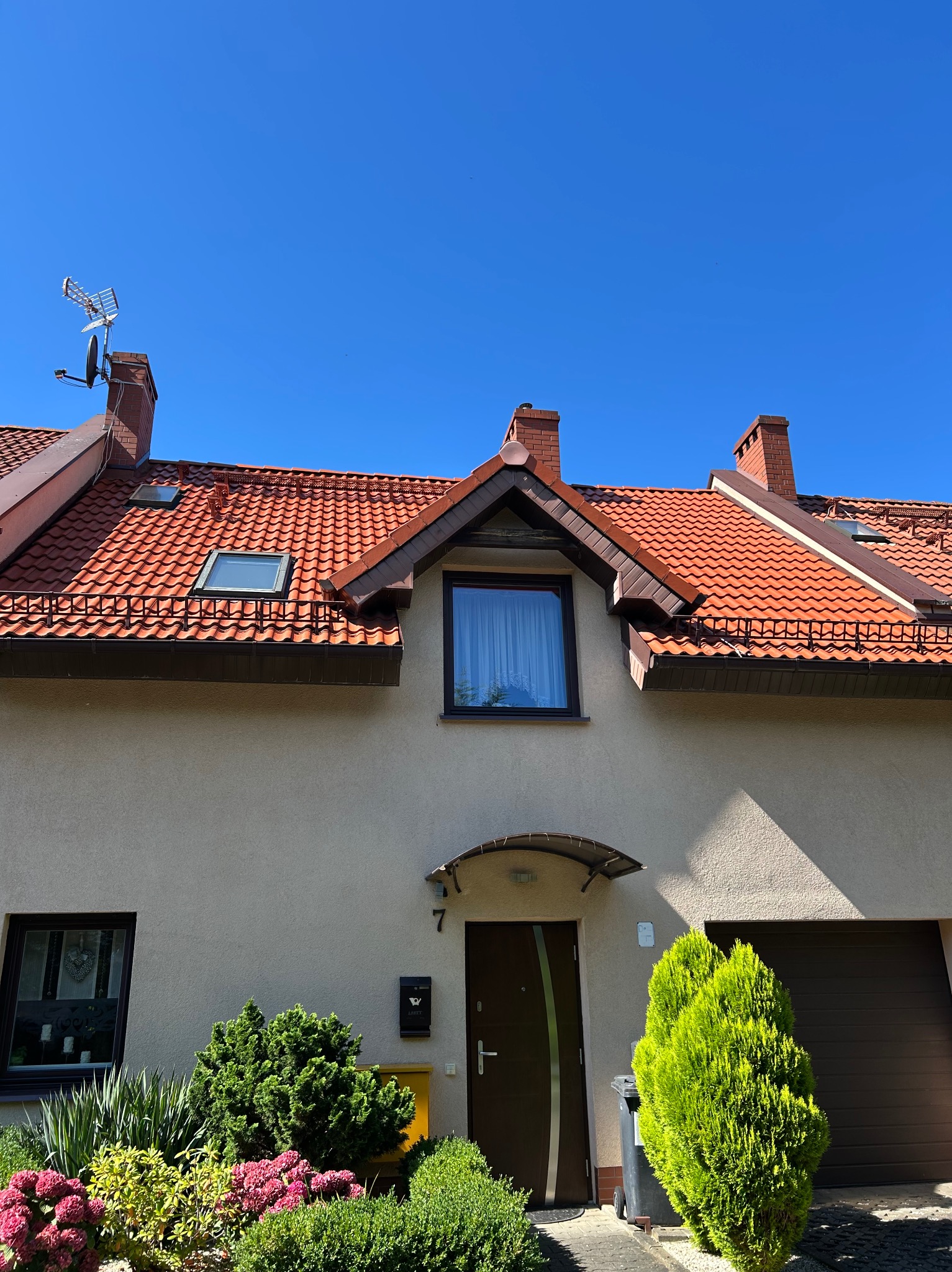 Dom jednorodzinny z czerwoną dachówką w słoneczny dzień. Widoczny fragment elewacji, okna, drzwi wejściowe i zadbany ogródek.