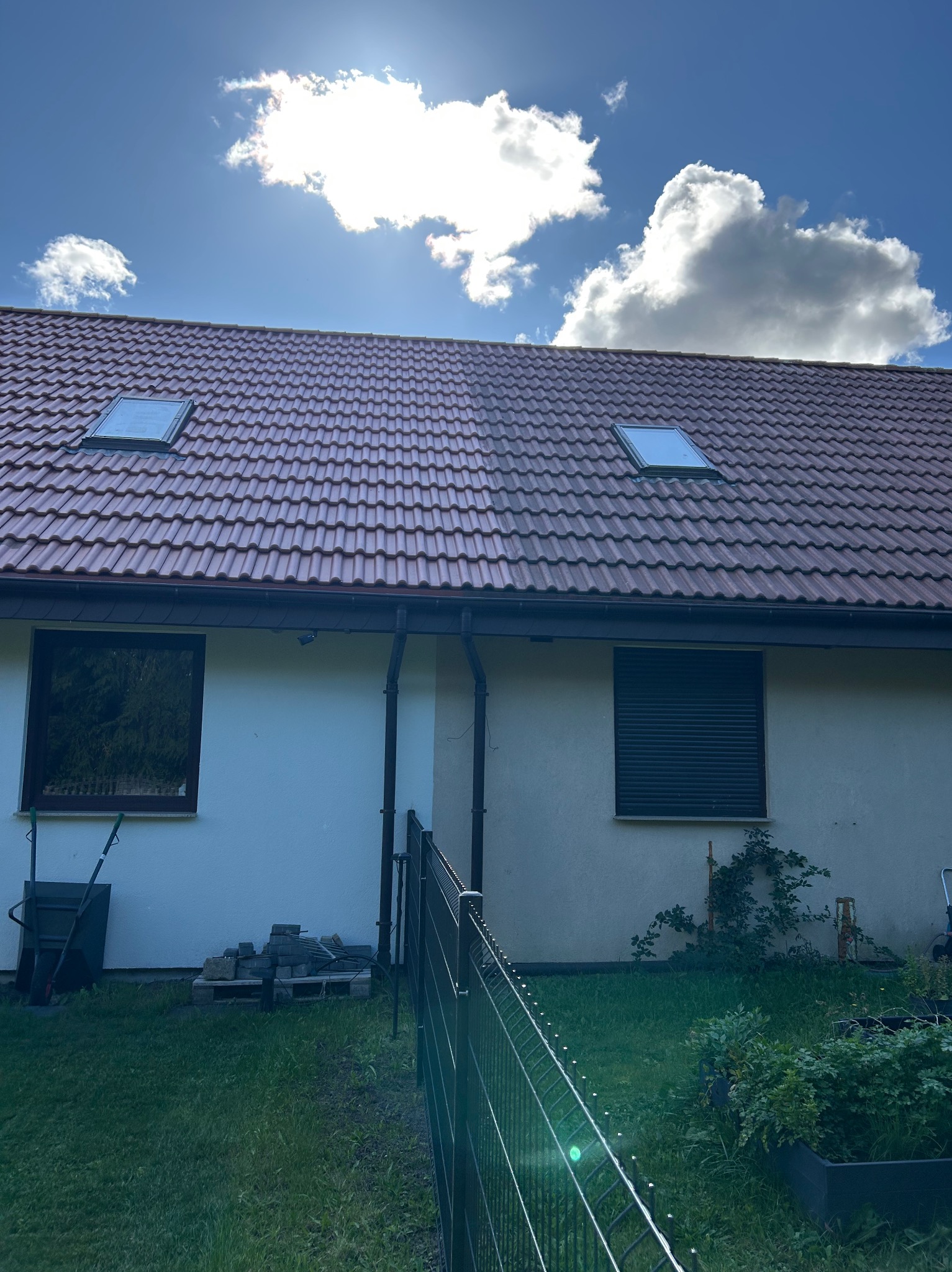 Dom z czerwoną dachówką i dwoma oknami dachowymi w słoneczny dzień. Widoczne rynny, fragment ogrodu z ogrodzeniem i narzędzia ogrodnicze przy ścianie.