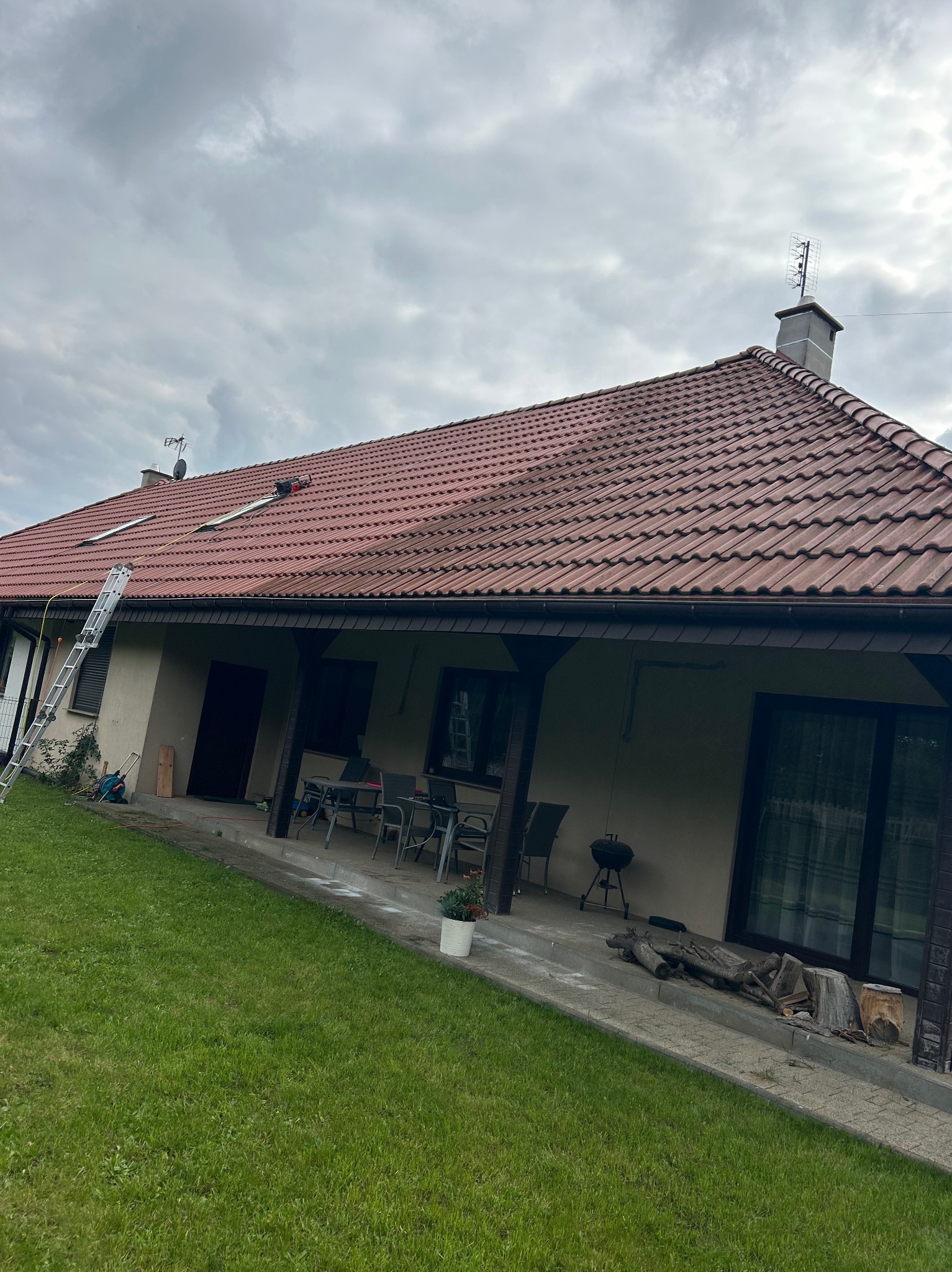 Ceglasty dach domu jednorodzinnego po czyszczeniu, z drabiną opartą o rynnę. Widoczne ślady czyszczenia na powierzchni dachówek, pochmurne niebo w tle.