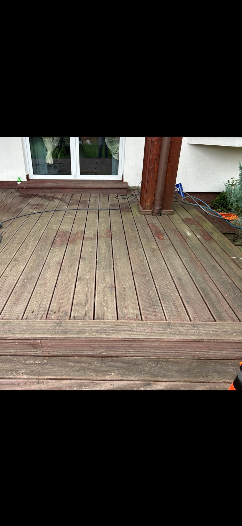 Drewniany taras z widocznymi śladami użytkowania, z elementami architektury ogrodowej i fragmentem elewacji budynku w tle. Widoczne zabrudzenia na deskach.