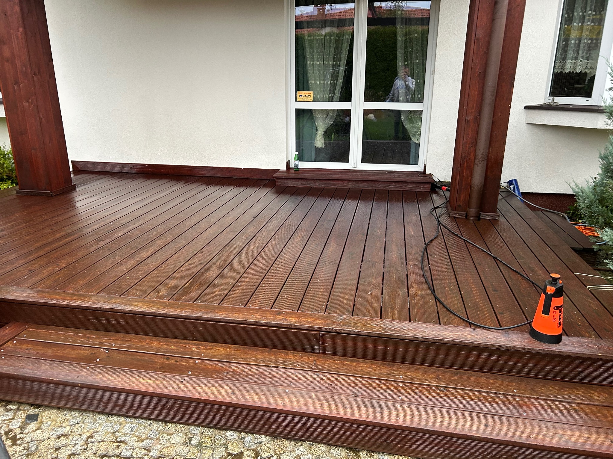 Impregnowany drewniany taras z widocznymi schodami i elementami konstrukcyjnymi. Na tarasie stoi opryskiwacz. W tle biała ściana budynku z oknem.