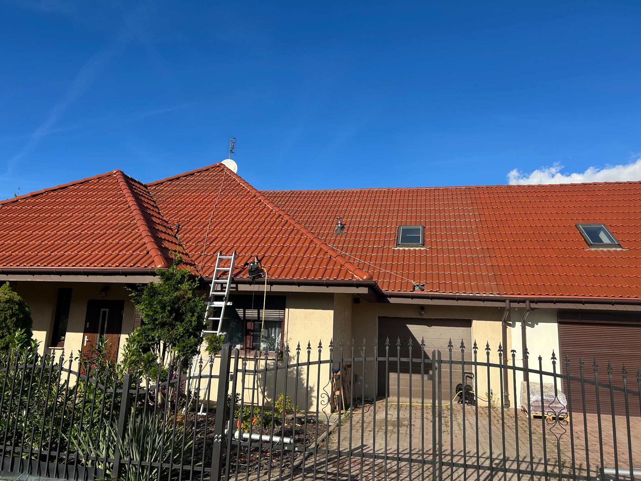Dom z czerwoną dachówką widziany zza metalowego ogrodzenia. Na dachu drabina. Widoczne okna dachowe i antena. Błękitne niebo w tle.