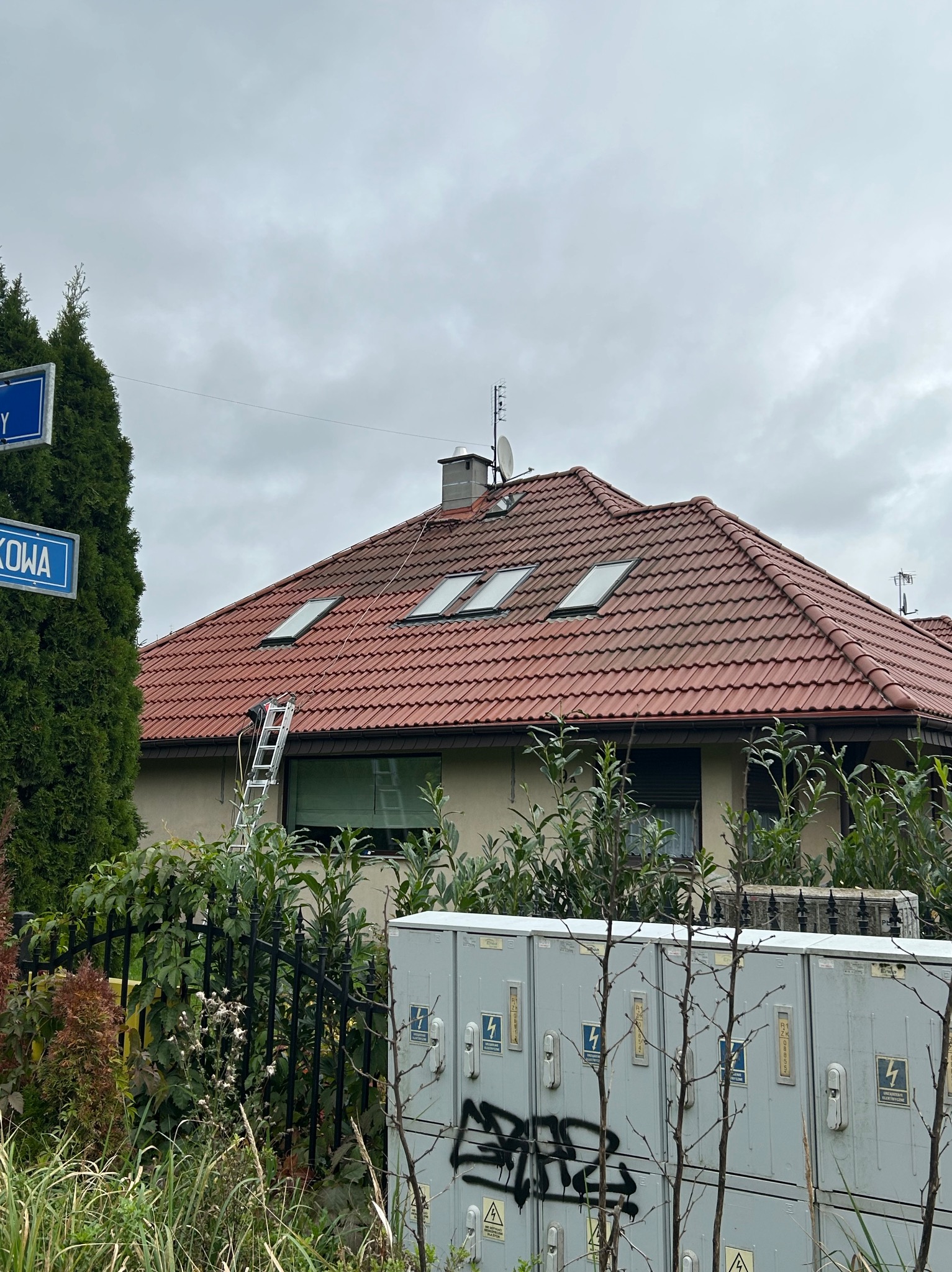 Czerwony dach domu z oknami dachowymi widziany z poziomu ulicy, drabina oparta o rynnę, pochmurne niebo w tle. Widoczne skrzynki elektryczne i roślinność.