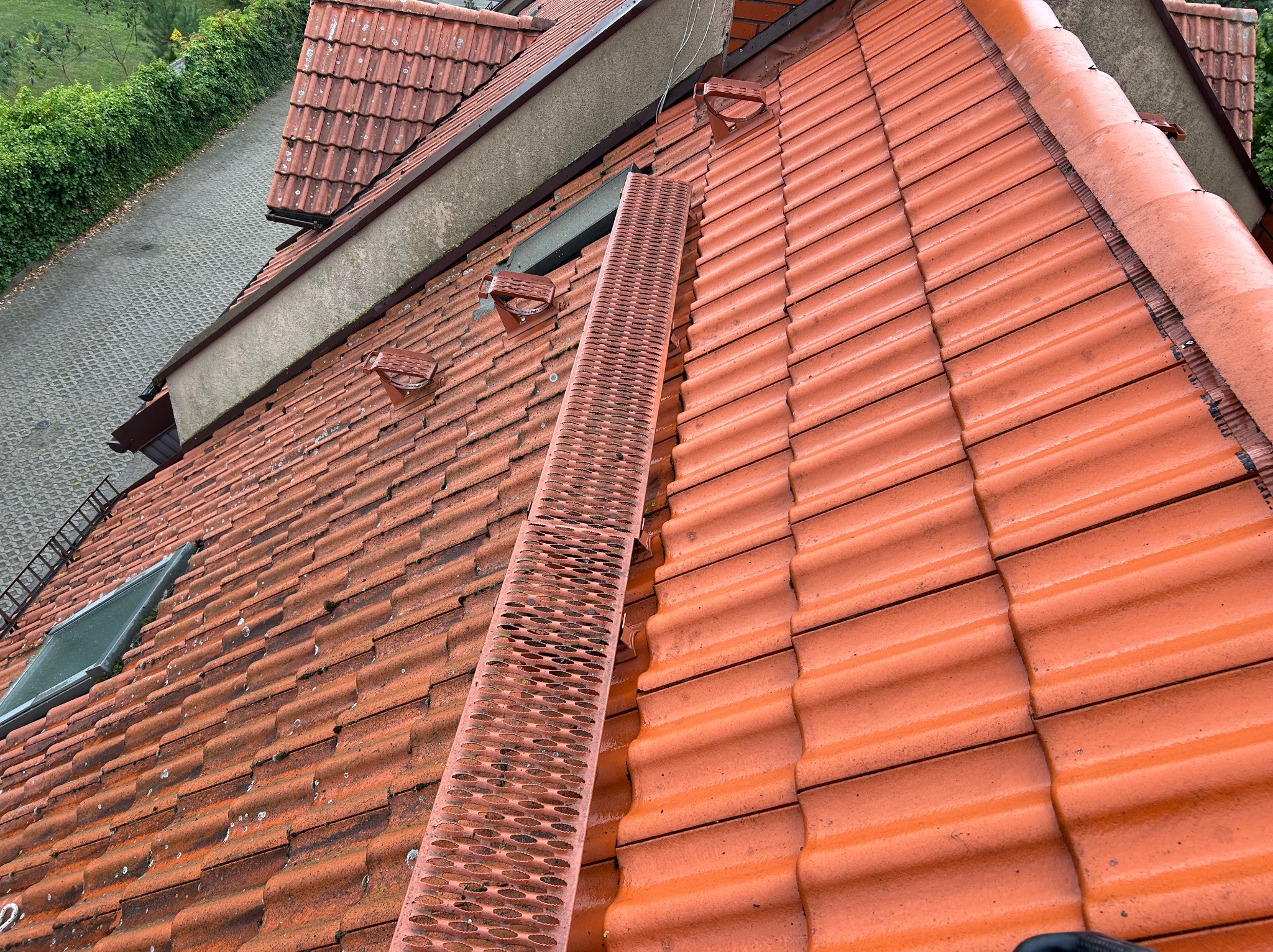 Czerwona dachówka z ławą kominiarską na dachu w Szczecinie. Widoczne okno dachowe i fragment chodnika z zielenią w tle. Ujęcie z góry.